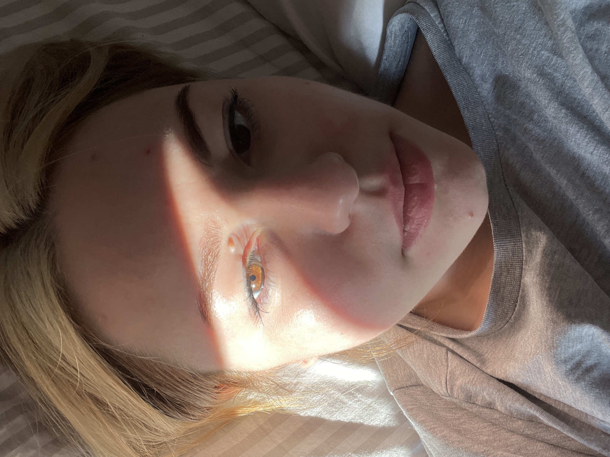 A fair-skinned woman with blonde hair lies on a striped bed in a room illuminated by sunlight. She is wearing a gray shirt. A beam of light diagonally bisects her face. Her eyes are open. No specific location is identifiable in the image. The absence of landmarks or recognizable features prevents the identification of the location as Boreti, Montenegro.