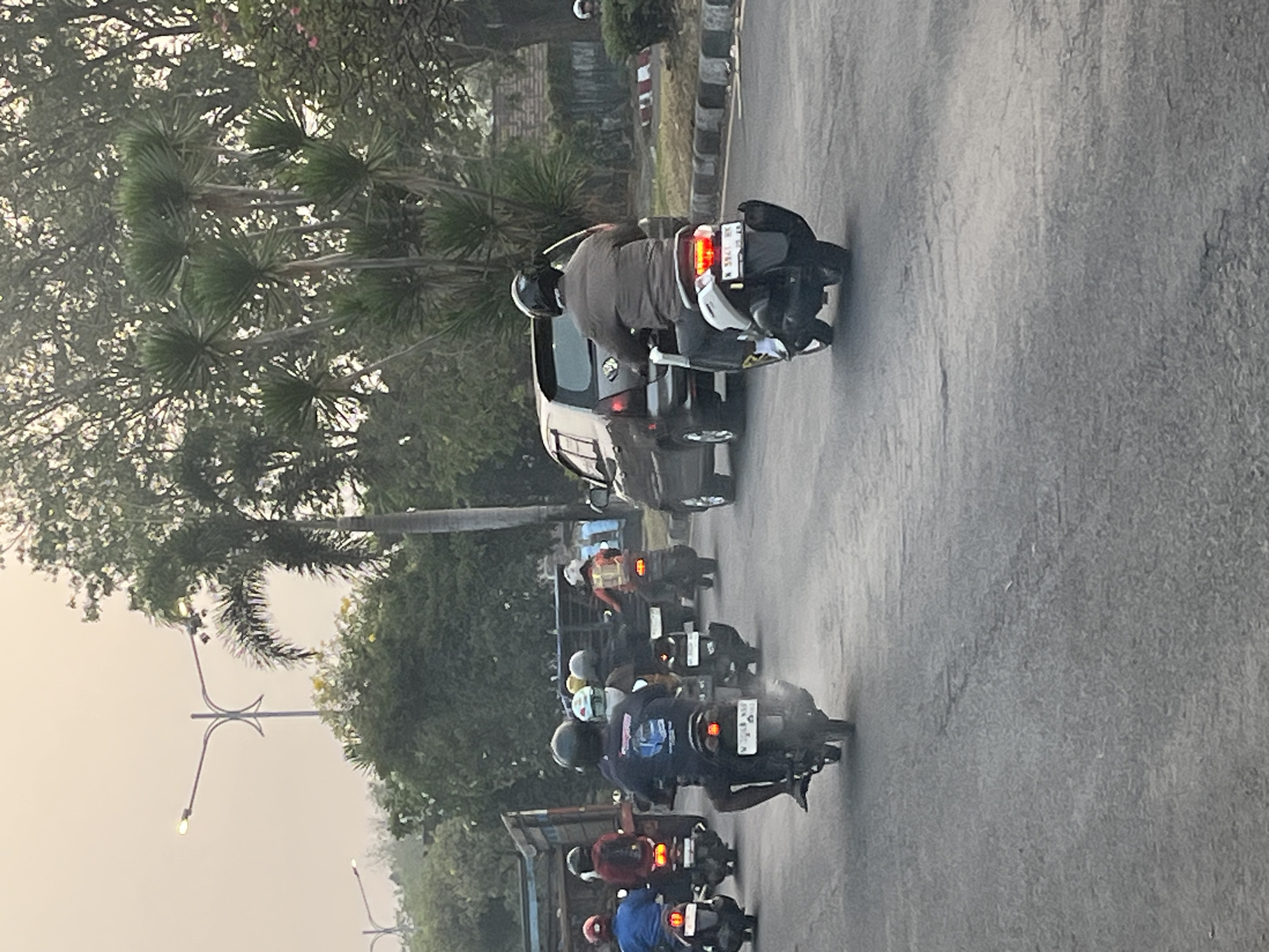The image shows a street scene with multiple motorcycles and a car on a road. Several motorcycles are moving in the same direction. One motorcycle in the foreground, on the right side of the image, has a rider wearing a brown jacket and dark pants, with a black helmet. The motorcycle is silver and black, with a license plate reading "N 3941 HK". To the left of this motorcycle, there is a dark-colored car. Further down the road, there are more motorcycles with riders. On the left side of the road, there are two trucks, one with a red cab and a wooden bed, and another with a blue bed. The road surface appears to be asphalt, with some cracks visible. Trees and foliage are present on the sides of the road. The sky is overcast, with a slight haze. The scene appears to be in a semi-urban or suburban area, possibly Kludan, Indonesia, given the presence of multiple motorcycles, trucks, and the overall environmental setting.