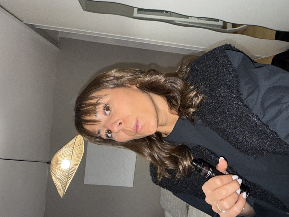 A woman with shoulder-length brown hair and bangs looks up with a slight pout. She wears a black t-shirt and dark textured sweater. In her right hand, she holds a vape pen. The room is a light gray. A decorative hanging light with a yellow shade is visible to the left of her. Behind her is a white square canvas hanging on the wall. The photo is tilted.