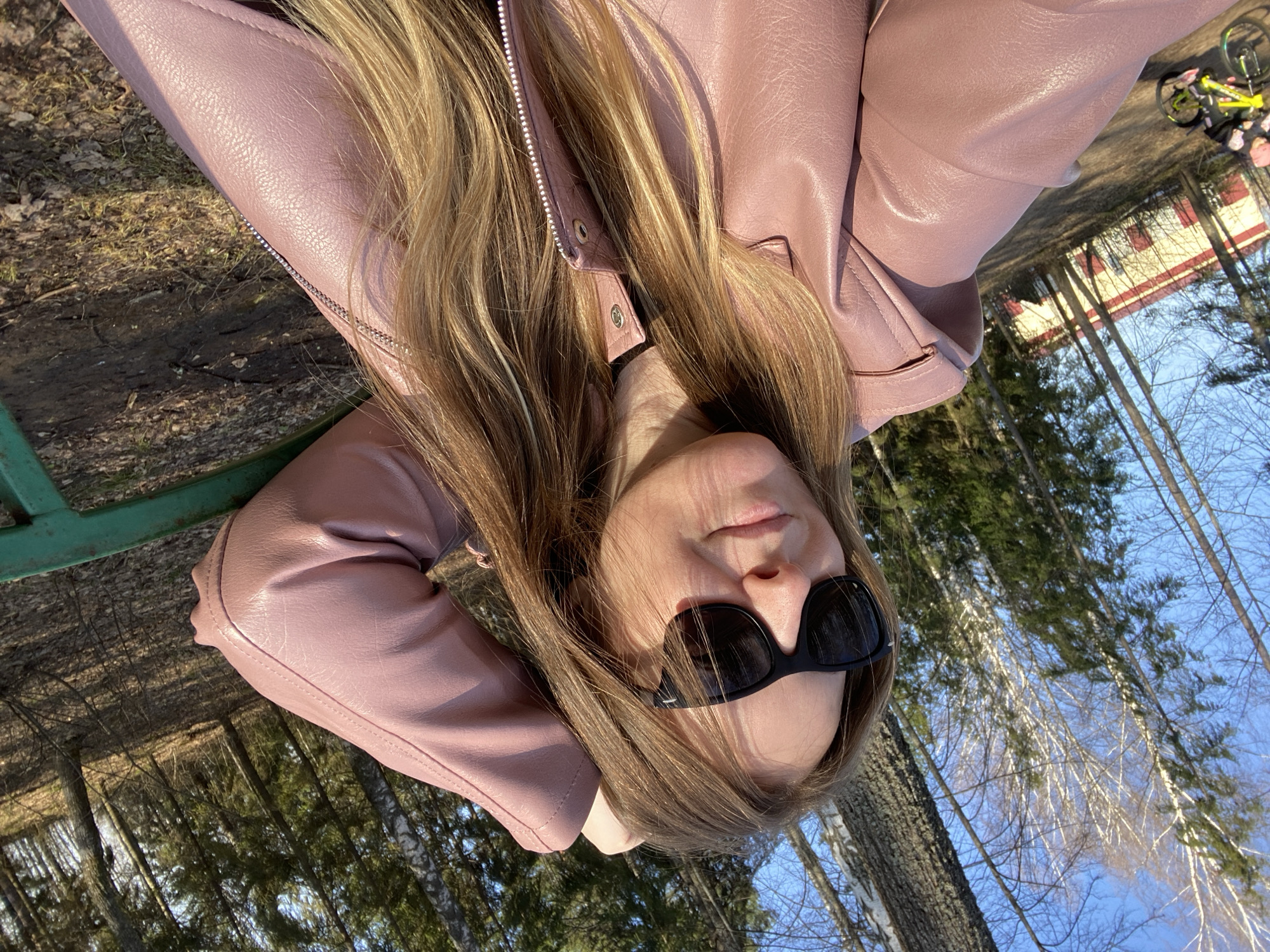 A young woman wearing a pink leather jacket and black sunglasses reclines on a green park bench in Noviy, Russia. Her long blonde hair falls around her face. The background shows trees, blue sky, and a building with red and white features. A yellow bicycle is visible in the distance. The image appears to be taken in early spring.
