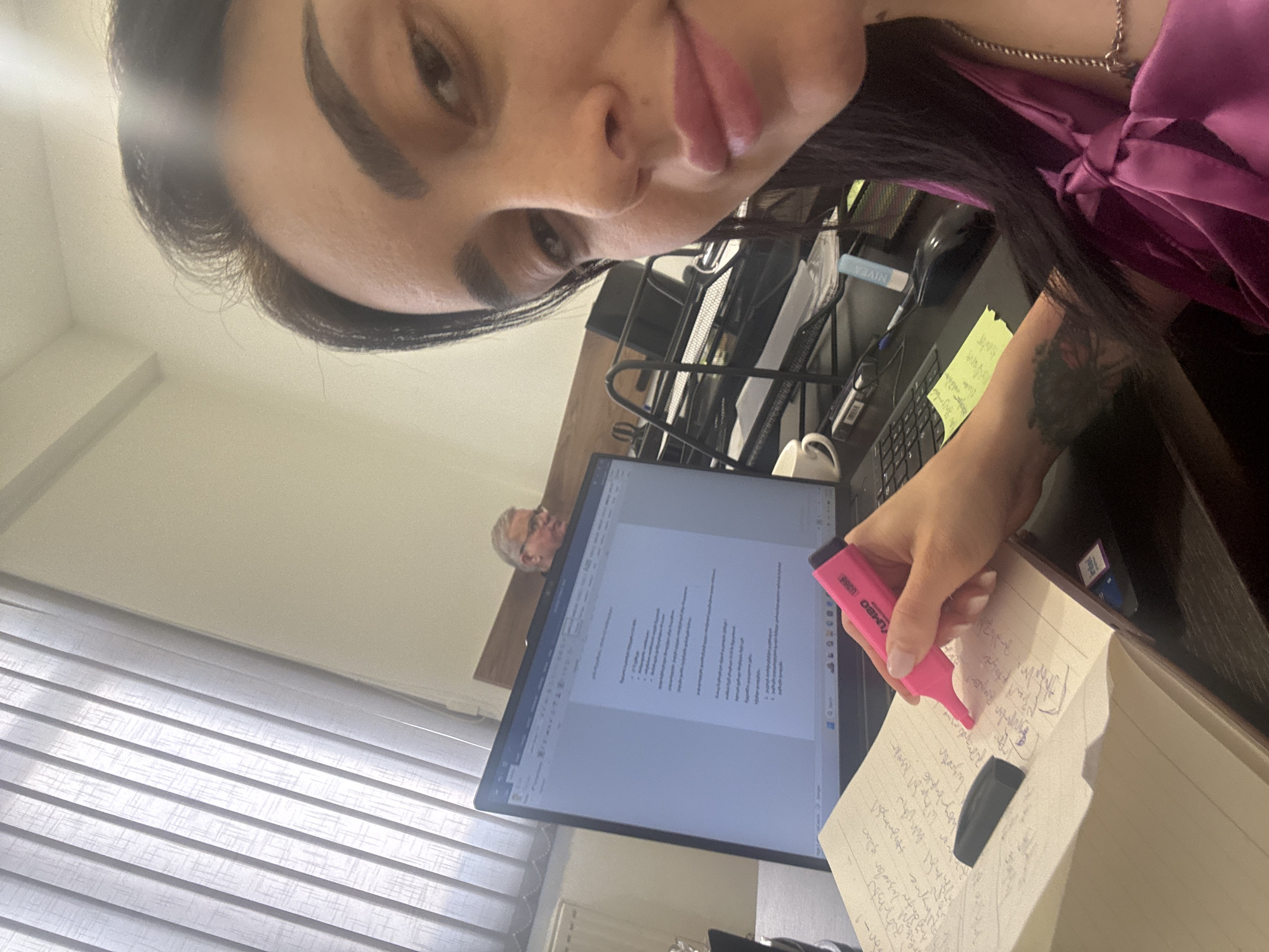 A woman is taking a selfie indoors. She is at a desk with a computer monitor in front of her displaying a document, and a notepad to the side which she is marking with a pink highlighter. There is another person visible on the monitor, possibly on a video call. The location is likely an office or home office in Tbilisi, Georgia, based on the window blinds and the general appearance of the room.
