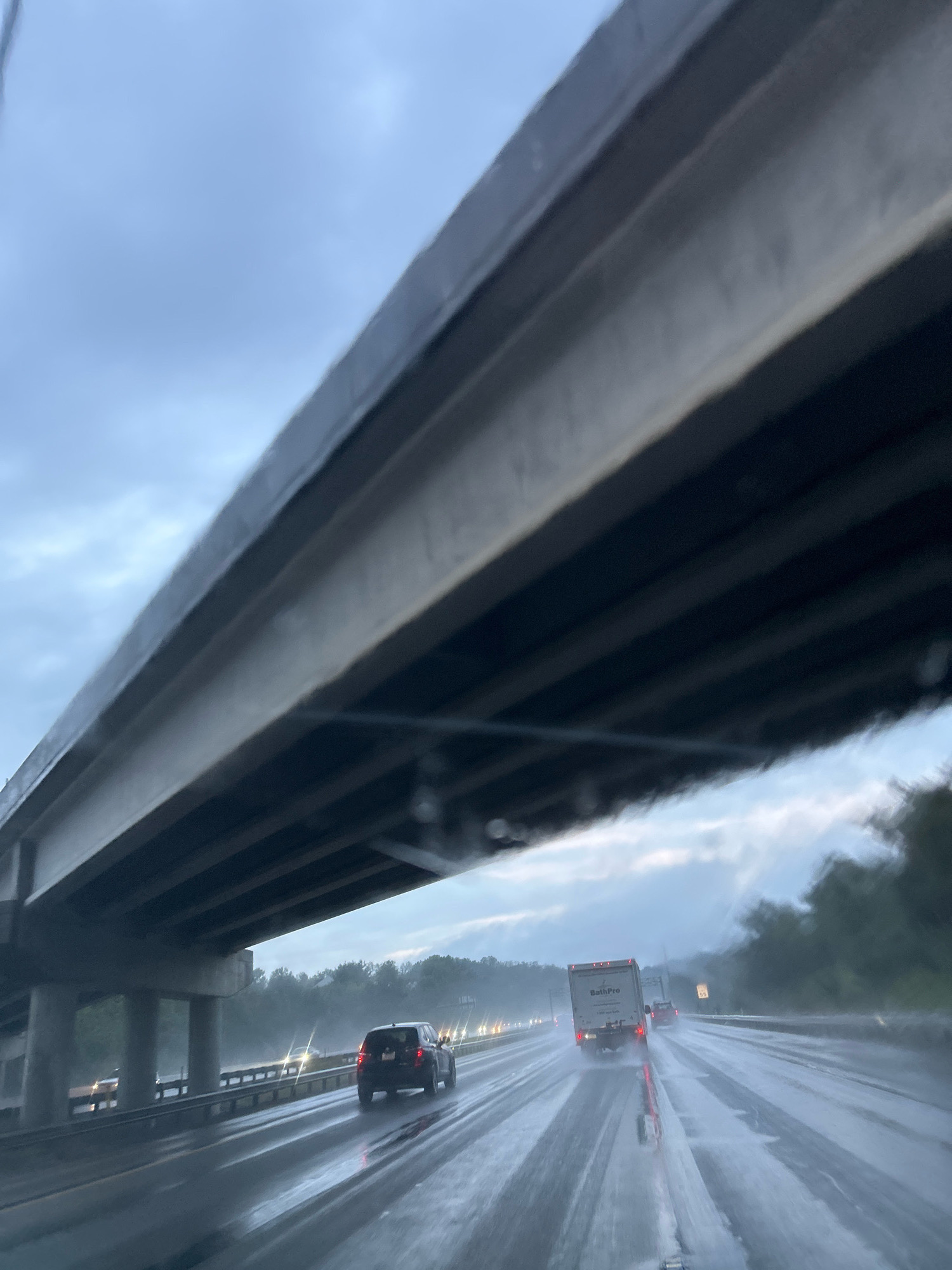 The image shows a view from inside a vehicle traveling on a wet highway in Collier Township, USA. A concrete overpass runs diagonally across the top of the frame, casting a shadow over the road. The highway is slick with water, with visible reflections and spray from the tires of passing vehicles. In the foreground, a dark SUV is visible in the right lane. Further ahead, a white box truck with the logo "BathPro" is in the left lane, followed by a red car. Distant taillights indicate more traffic. The sky is overcast and appears to be either dawn or dusk. Trees line the side of the highway in the distance.