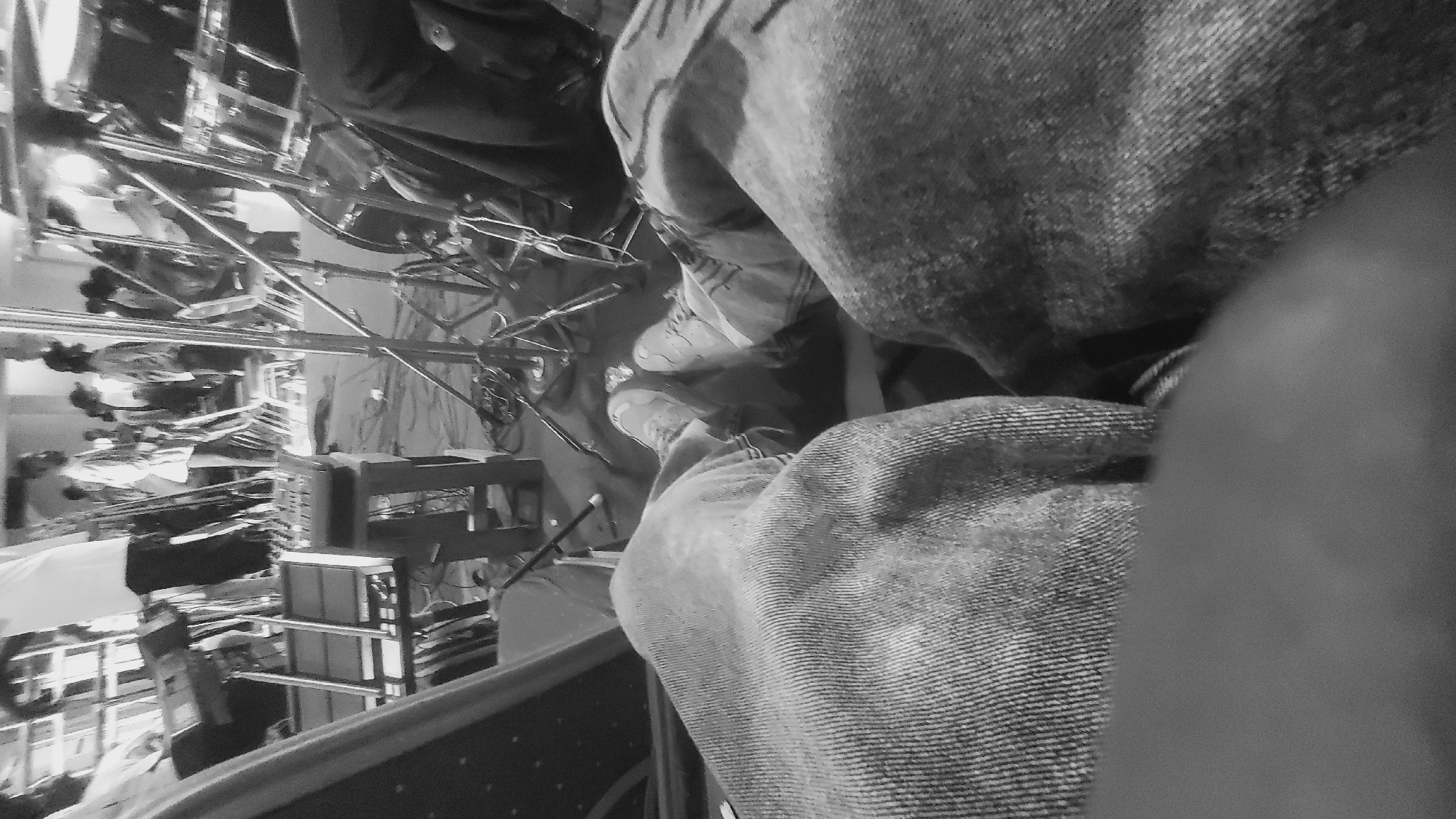 A low-angle, black and white, first-person perspective shot shows the legs of an individual wearing denim jeans and light-colored sneakers. The setting appears to be a performance or event space, possibly in Jalingo, Nigeria, with stage equipment and other individuals visible in the background.
In the foreground, the denim-covered thighs and knees of the photographer dominate the frame. To the right, the lower legs and feet, clad in sneakers, are also visible, suggesting the photographer is seated or standing with their legs spread. The texture of the denim is prominent, showing the weave of the fabric.
The background is a blur of activity and equipment. To the left, a keyboard or synthesizer is set up on a stand. Further back, several people are standing, some appearing to be performers or crew members, dressed in what looks like casual attire. Metallic structures, likely part of the stage rigging or lighting equipment, are scattered throughout the midground. A drum set can be seen to the right. The floor appears to be covered in a dark material, possibly a stage mat or tarp, with wires and cables strewn across it. The overall impression is a behind-the-scenes or audience perspective of a live event.