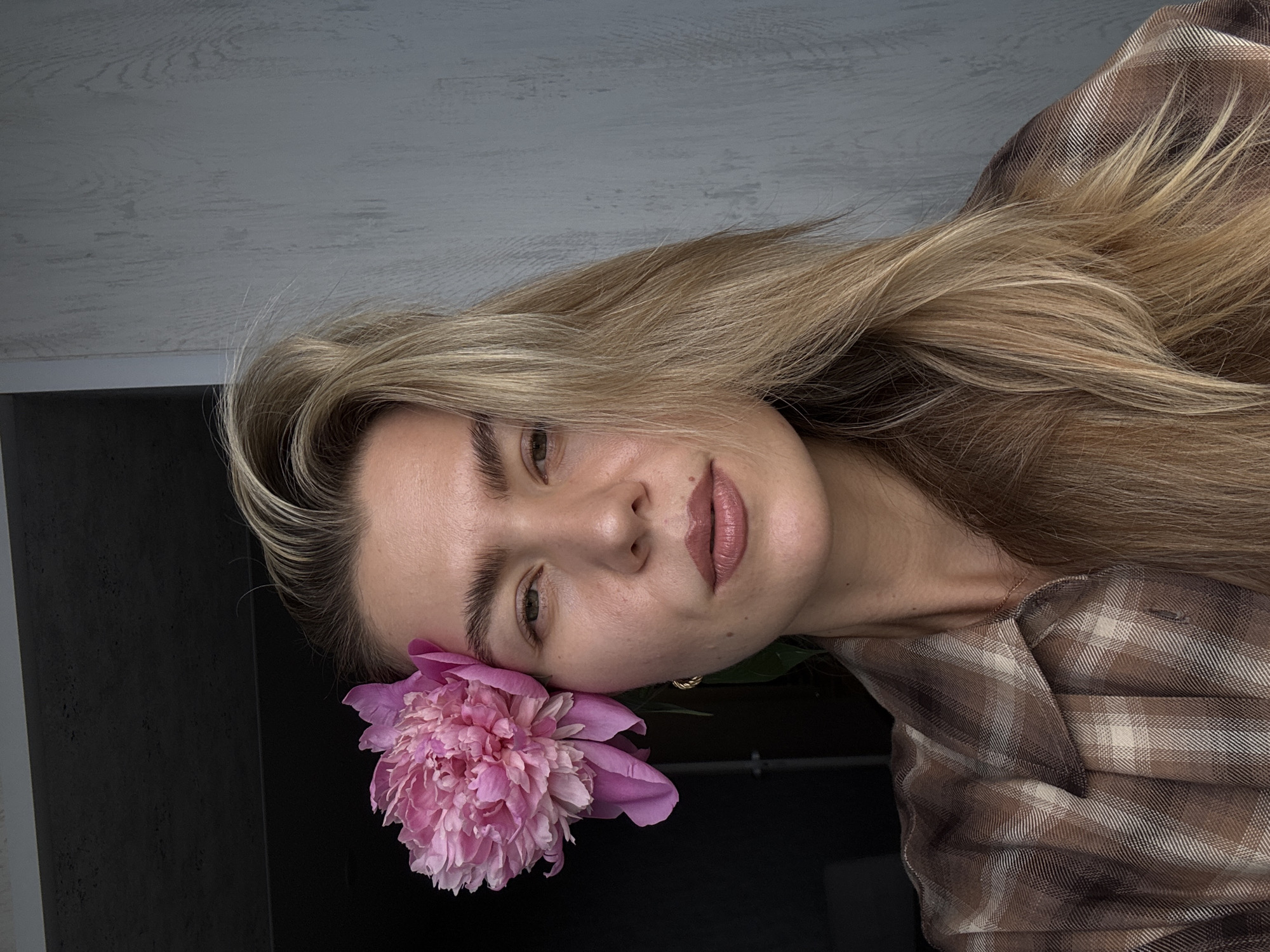 A head-and-shoulders portrait depicts a woman looking directly at the camera. She has long, light brown hair parted to the left and falling over her right shoulder. A large, vibrant pink peony flower is positioned behind her left ear, resting against her temple. Her eyebrows are dark and prominent, and she has light-colored eyes. Her lips are full and painted with a matte, nude-pink lipstick. A small gold hoop earring is visible in her left ear. She is wearing a brown and beige plaid collared shirt. The background on the left is dark and indistinct, while the background on the right is a light grey wall with a subtle vertical wood grain texture.