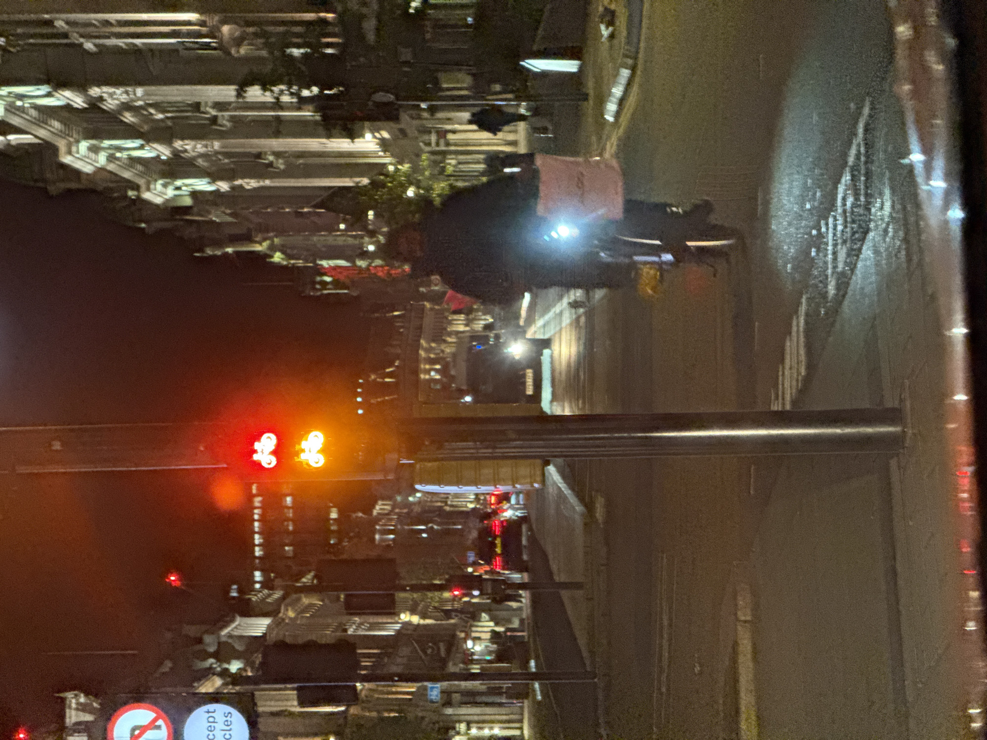 The image depicts a nighttime street scene in London, United Kingdom. A cyclist with a front light and a bag on their back is riding on the right side of the road. The street is wet, reflecting light from street lamps and vehicles. A traffic light displaying both red and amber symbols for cyclists is visible. Street signs indicating restrictions for cyclists are also present. Buildings line the left side of the street, illuminated by street lights. Cars are visible further down the street. The image appears to have been taken from inside a vehicle.