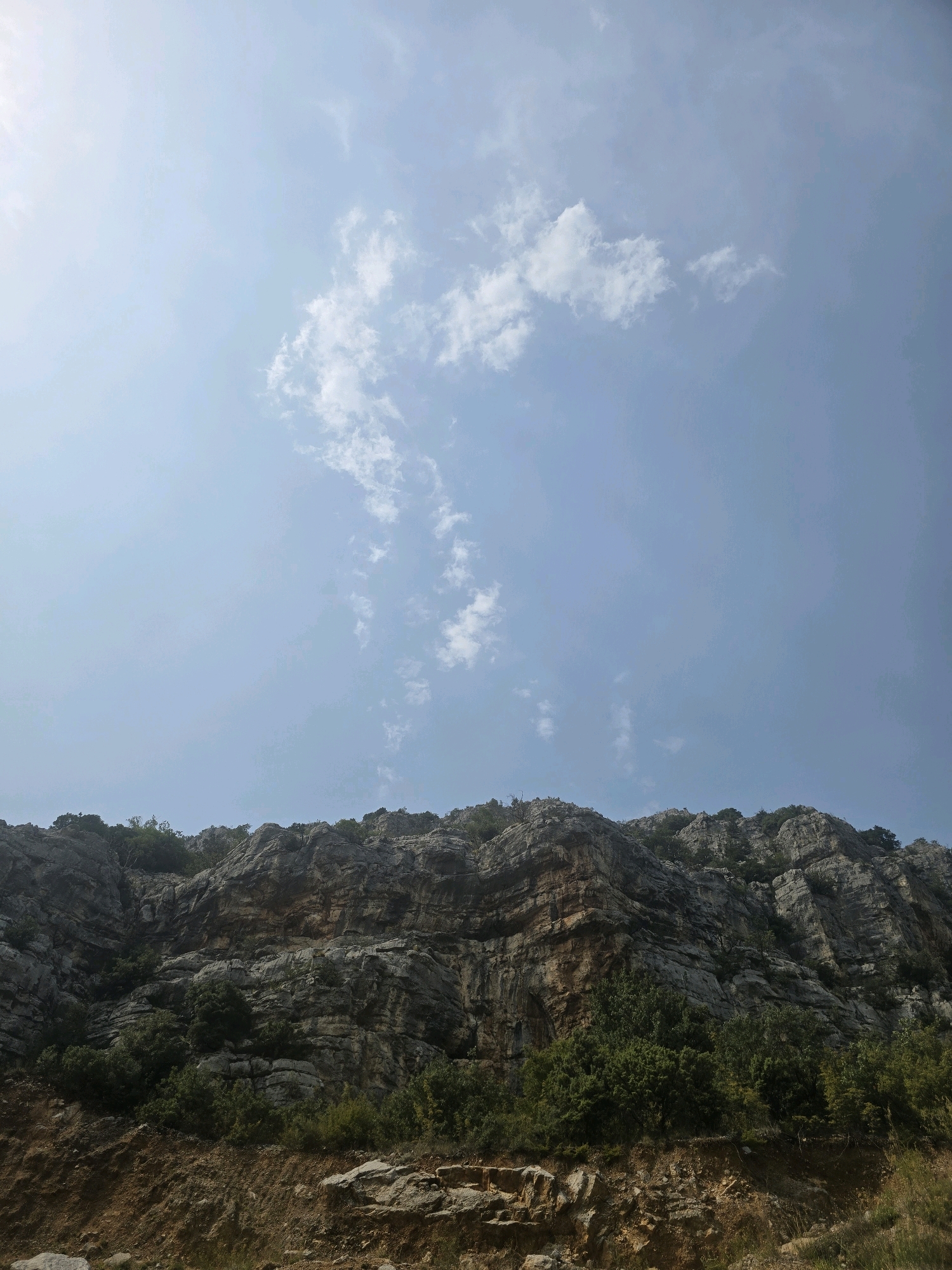 The image depicts a steep, rocky mountainside with sparse green vegetation growing from its crevices and along its base. The rocks are varied in color, ranging from grey and brown to some reddish hues, indicating stratification and erosion. The terrain at the bottom of the image appears to be a dirt road or embankment, with loose stones and soil. The sky above is a pale blue with scattered white clouds, suggesting a daytime scene. The overall impression is one of a rugged, natural landscape, likely in a mountainous region. The location is identified as being near the city of Podgorica, Montenegro.
