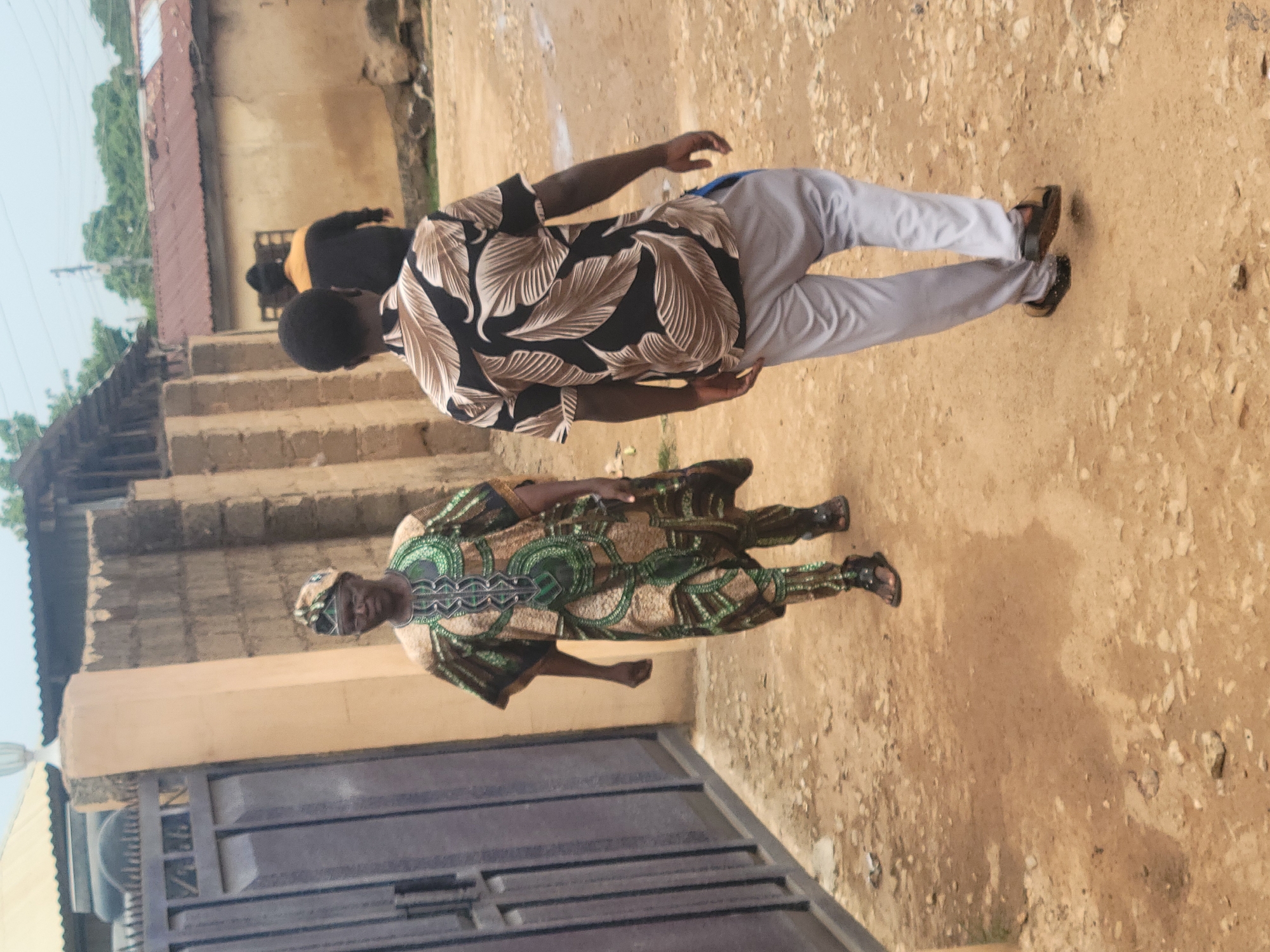 The image depicts an outdoor scene in Jos, Nigeria. Two men are prominently featured: One is wearing a patterned, green and brown traditional outfit, while the other is wearing a dark shirt with leaf-like patterns and light gray pants. They are standing on a dirt ground. A building with a metal gate is visible on the left, and in the background, there is a building with steps and other people on the steps.