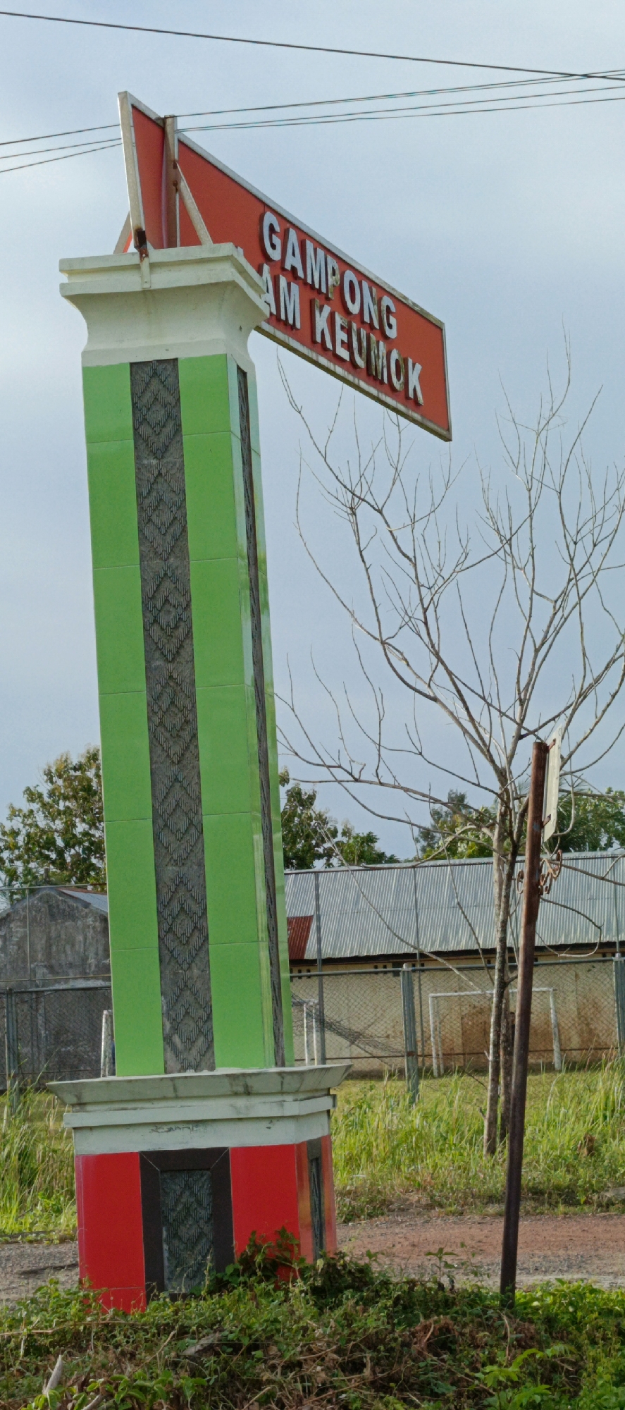 An outdoor, eye-level shot captures a monument sign at the entrance to Gampong Lam Keumok, Indonesia. The sign is a tall, slender structure with a red rectangular top adorned with white text that reads "GAMPONG AM KEUMOK". The main body of the monument is green with a decorative patterned inlay. The base of the monument is red with dark patterned inlays. The monument stands on a patch of overgrown greenery and dirt. In the background, a chain-link fence encloses an area with buildings that have corrugated metal roofs. A leafless tree with thin branches stands to the right of the monument. The sky is overcast and pale blue. No people are visible in the frame.