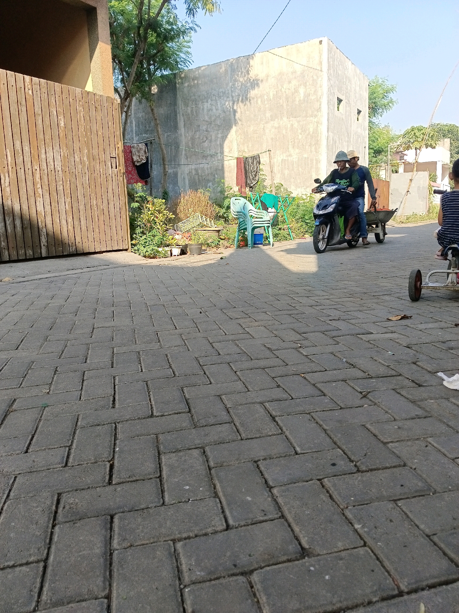 In Krembangan, Indonesia, two men are riding a motorcycle, with one man driving and the other sitting behind him, holding onto the driver. They are pulling a small cart with what appears to be red material inside. The motorcycle is dark blue and green. The men are wearing casual clothing and hats. The scene is set on a paved street, with a wooden fence on the left and a concrete building in the background. There are also some plants and clothes drying on a line in the background. A child is visible in the distance, sitting on a small toy vehicle. The lighting suggests it is daytime.