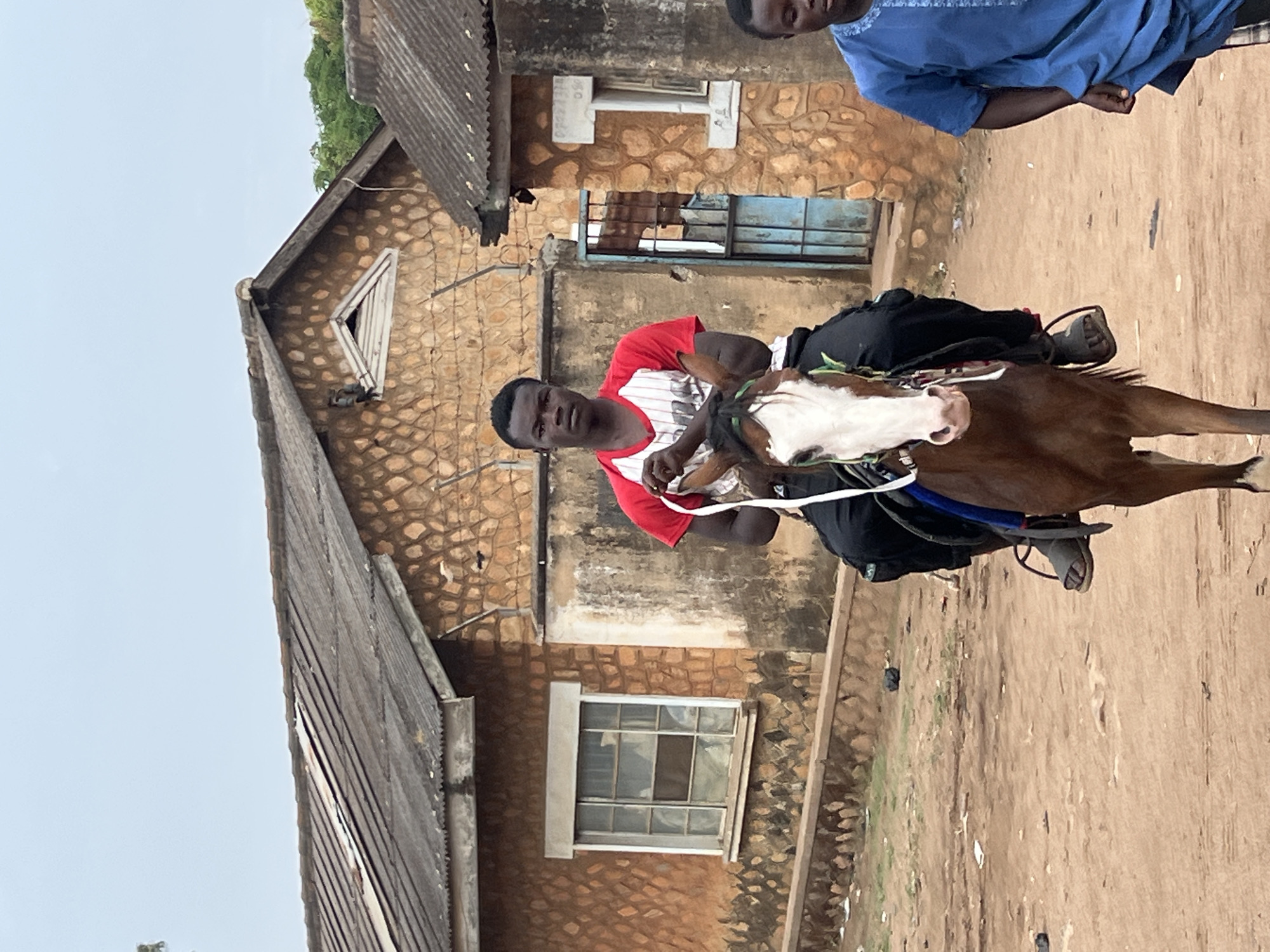A medium outdoor shot depicts a man riding a horse in Bukuru, Nigeria. The man, wearing a red and white t-shirt, is seated on a brown and white horse. He is holding onto the reins. The horse has a blue saddle and is standing on a dirt ground. In the background is a building with a stone facade and a corrugated metal roof. There are two windows visible on the building. Another man wearing a blue shirt is seen partially in the upper right corner of the image. The sky is overcast. The overall scene suggests a rural or semi-rural environment.