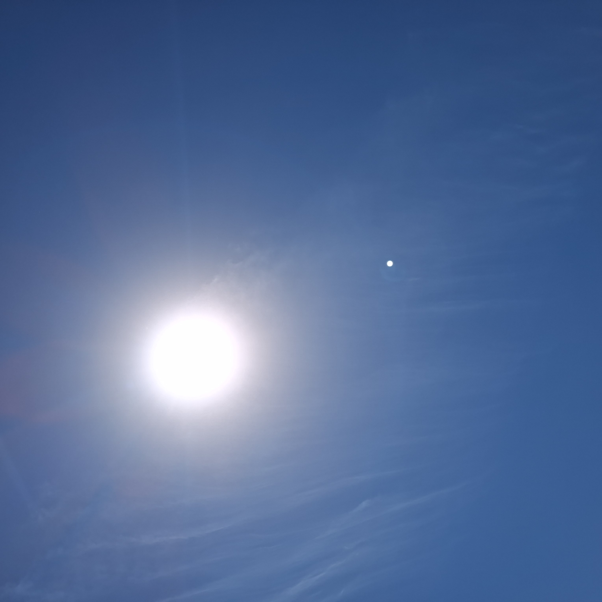 The image shows a bright sun in a blue sky with some wispy clouds. A bright white spot is also visible in the sky to the right of the sun. The location is unknown.