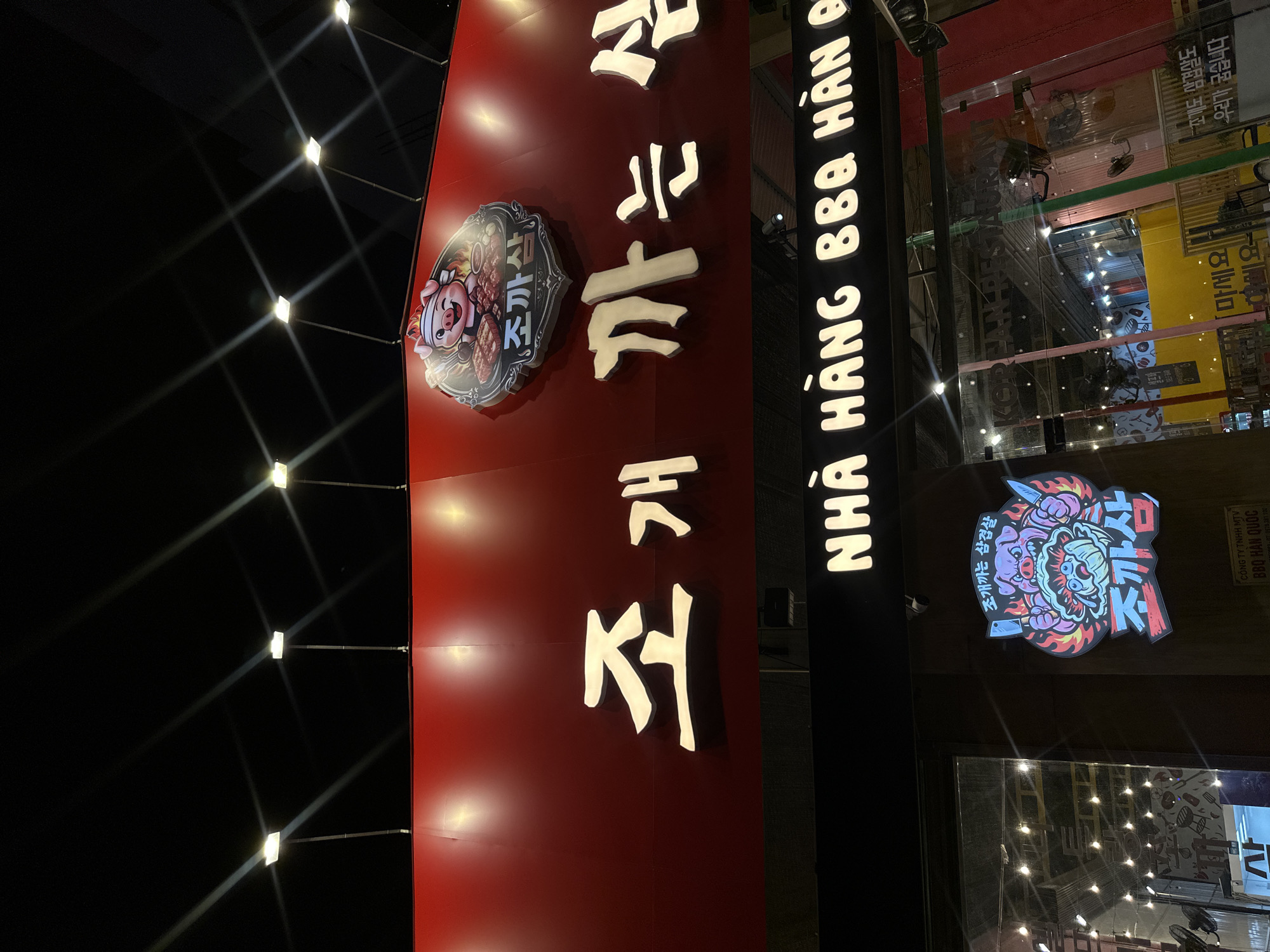 The image displays the illuminated facade of a Korean BBQ restaurant at night, located in Nha Trang Ward, Vietnam. The scene is characterized by bright signage against a dark sky.
A prominent red sign on the left features large, white Korean characters, reading "조까삼" (Jjokkassam) and "조개 까는 삼겹살" (Jogaekkaneun Samgyeopsal), which translates to "Clam-peeling Pork Belly." This sign also includes a cartoon pig logo holding grilling utensils. Multiple square-shaped lights on poles cast bright beams across the red sign and into the dark night.
Adjacent to the red sign, a vertical black sign with glowing white letters spells out "NHÀ HÀNG BBQ HÀN B," indicating a "Korean BBQ Restaurant." A small surveillance camera is mounted on this sign. Below it, another sign features a colorful, stylized demon-like figure holding knives, again displaying "조까삼" and "조개까는 삼겹살" in Korean.
The building behind the signs has large glass windows, reflecting interior lights and additional textual information. Reflected Korean text visible includes "조개도 삼겹살도 우리가 굽습니다" (We grill both clams and pork belly), further detailing the restaurant's offerings. Other visible text snippets include "CÔNG TY TNHH MTV BBQ HAN QUOC" and "마세요". The setting suggests a lively commercial establishment operating after dark. No people are visible in the image.