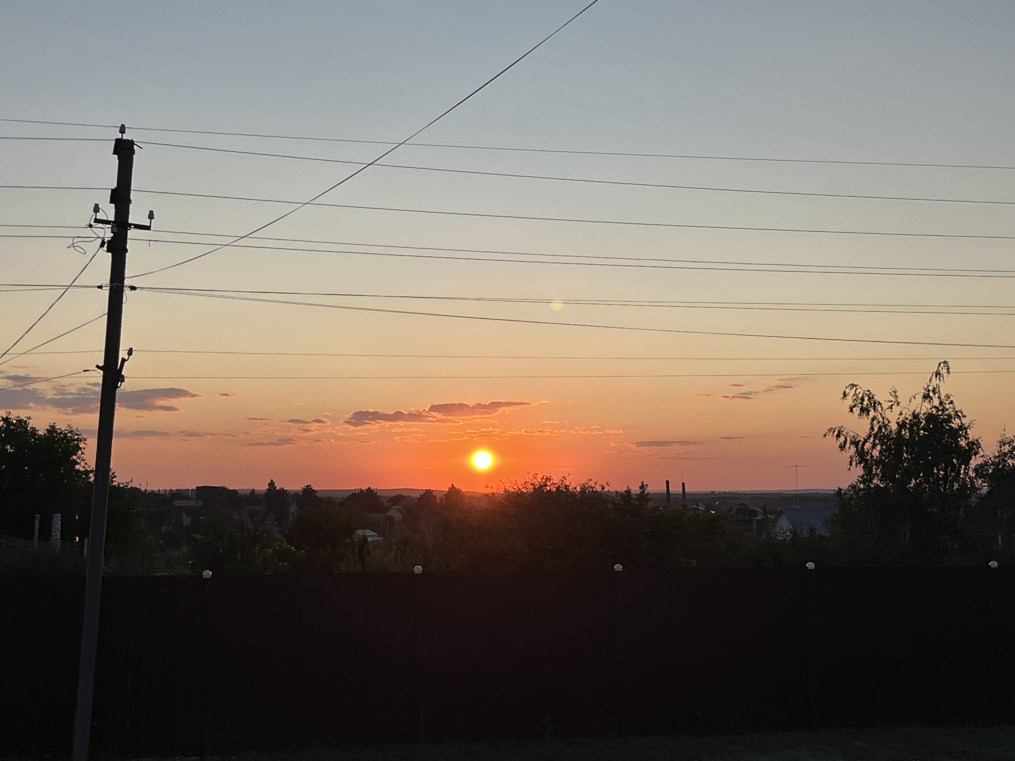 This is a photograph of a sunset over a rural landscape. The sun, a bright orange orb, is low in the sky, casting a warm glow over the scene. Clouds are scattered across the sky, some tinged with pink and orange. In the foreground, a dark fence runs horizontally across the image, obscuring the lower portion of the landscape. Behind the fence, trees and buildings are visible, silhouetted against the colorful sky. A telephone pole with wires stands on the left side of the image, adding a man-made element to the natural scene. The overall impression is one of a peaceful evening in a rural setting. The location is believed to be in the outskirts of Saratov, Russia, given the architectural style of the visible buildings and the surrounding vegetation. No people are present in the image.