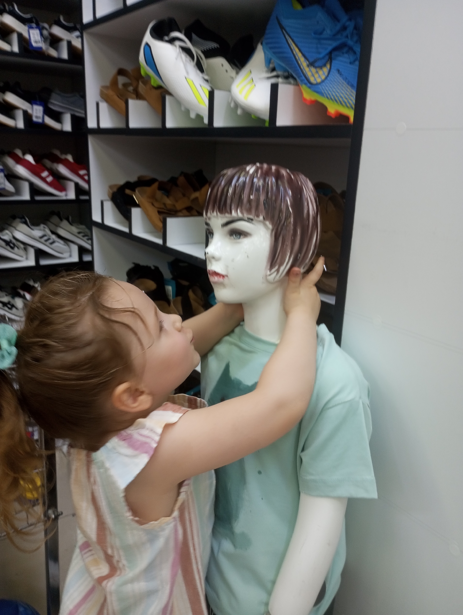 The image shows a young child with light brown hair, wearing a striped dress, embracing a mannequin. The mannequin has short brown hair and a light blue t-shirt. The child is pressing their face against the mannequin's neck. In the background, shelves are filled with shoes, including athletic shoes, sandals, and what appear to be sneakers. The shelves are white and organized, with some shoes displayed on top and others within compartments. The overall setting appears to be a shoe store. The lighting suggests an indoor environment, possibly during the day given the brightness. The child's expression and embrace convey affection or playfulness. No text is clearly visible in the image. The location context of Tbilisi, Georgia is provided.