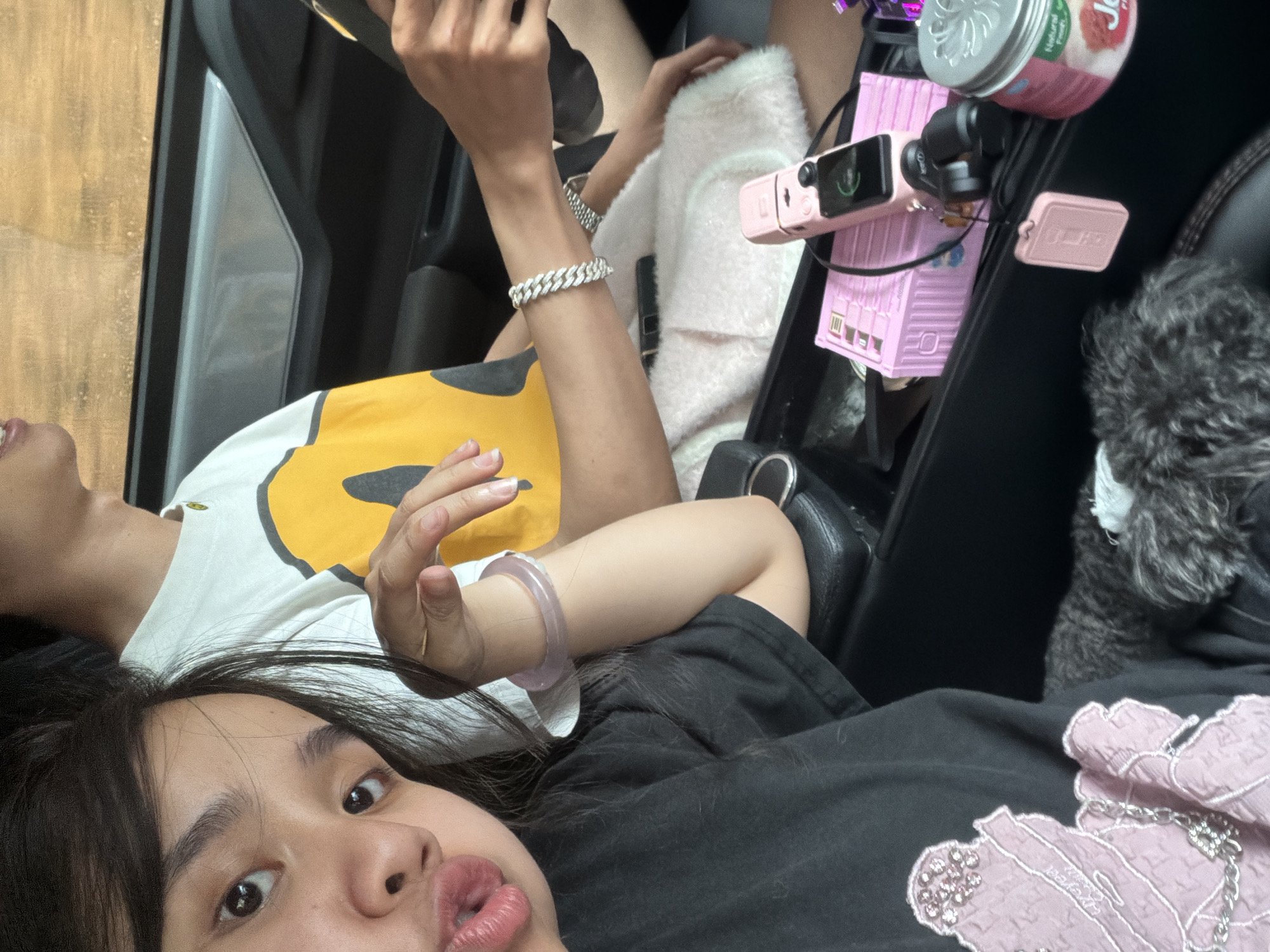 The image captures a casual selfie-style moment inside a vehicle, likely a car, in Măng Đen, Vietnam. The shot is angled, looking obliquely downwards from the lower left towards the upper right of the car's interior.
In the foreground, a person with long dark hair looks directly at the camera, featuring wide eyes and slightly parted lips, conveying a playful or surprised expression. They are wearing a black top embellished with a textured pink design on the right shoulder, which includes a silver chain accessory and faintly visible "Hypert" text. A light purple or lavender bangle adorns their left wrist.
Behind this individual, partially visible, is a second person, likely seated in the car's passenger seat. This person is wearing a white t-shirt with a prominent yellow and black graphic, possibly a stylized smiley face. They wear a silver Cuban link bracelet on their right wrist and a silver watch on their left. One of their hands is visible, holding a smartphone, suggesting they might be occupied with it.
The car's interior is primarily black. In the console area between the two occupants, several items are neatly placed. These include a pink DJI Osmo Pocket camera, a pink rectangular container with a textured, brick-like pattern, and a silver can with a pink label bearing the words "Jelly" and "Natural Fruit." Another small, pink rectangular object, possibly a USB drive or compact power bank, is also visible. To the far right, a fluffy, dark gray object, potentially a small dog or a furry accessory, is seen on the seat or console.
The background outside the window is out of focus, showing a warm, light brown textured surface, resembling wood. The scene is brightly lit, indicating daytime, and suggests a relaxed atmosphere.