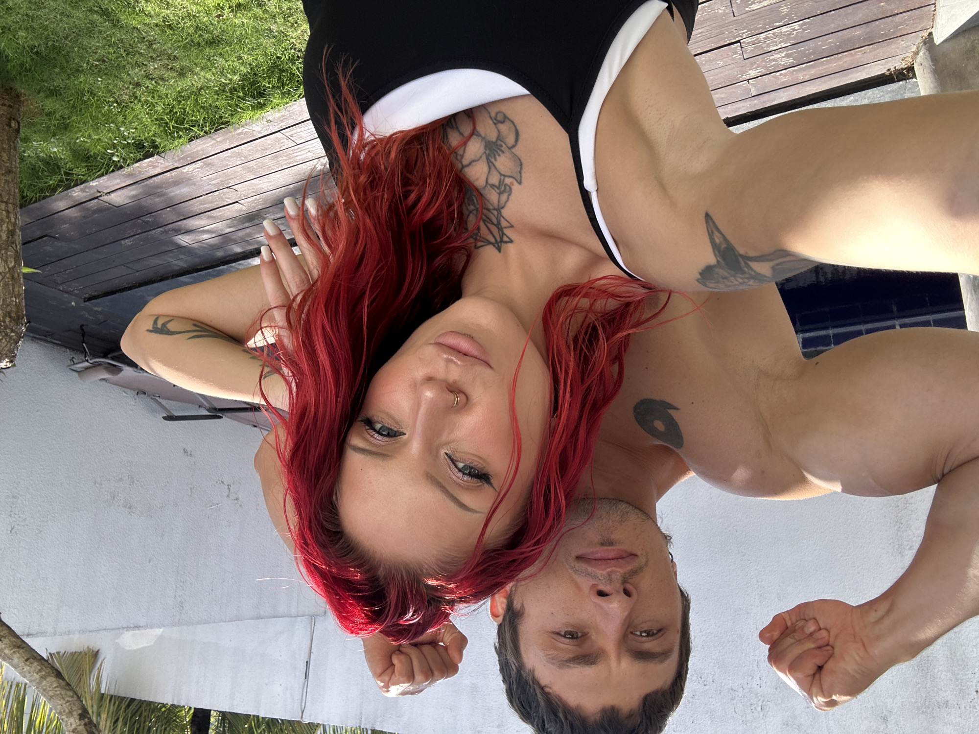 A man and a woman are posing for a selfie outdoors. The man, shirtless and with a tattoo on his left shoulder, flexes his bicep with his right arm. The woman, with bright red hair, is in the foreground, also looking towards the camera. She is wearing a black and white tank top and has tattoos on her chest and arm. They are standing on a wooden deck, with lush green grass and palm trees visible in the background. The scene appears to be a residential area or resort in North Kuta, Indonesia, given the tropical foliage and bright sunlight. A pool can be seen to the left of the frame, and lounge chairs are visible behind the couple. The overall atmosphere is relaxed and casual, suggesting a vacation or leisure setting.