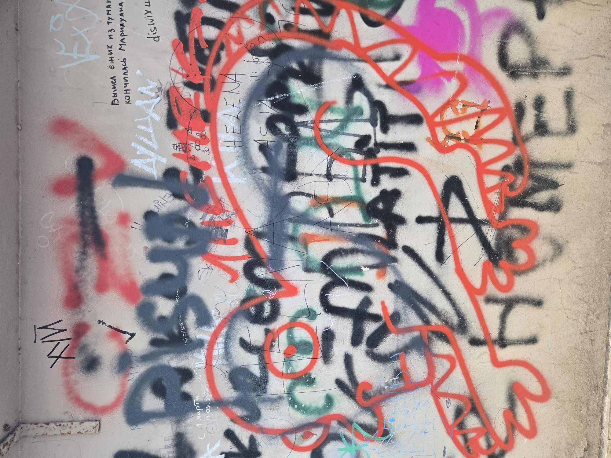 This image captures a heavily graffitied wall in Budva, Montenegro, filled with multiple layers of spray paint and marker text, primarily in red, black, and white. The wall itself appears to be a light-colored concrete or stucco, showing some texture and minor wear.
The most prominent feature is a large, stylized outline in bright red spray paint, forming what looks like an abstract cartoon character or creature with a prominent head, sharp teeth-like elements, and an eye-like swirl. This red artwork largely overlays other graffiti.
Beneath and around this red figure are numerous black spray paint tags, varying in thickness and style, some appearing as bold block letters and others as thinner, more intricate scribbles. White spray paint is also present, used for text and some outlines, and hints of pink and green spray paint are visible beneath the other layers, indicating the history of various additions. Fine-tipped marker or pen writings in black and blue further contribute to the dense overlay of markings.
Notable visible text includes:
- "С 1 марта" (From March 1st) in faint white.
- A Russian couplet in black pen: "Вышел ёжик из тумана Кончилась Марихуана." (A hedgehog came out of the fog. The marijuana ran out.), a humorous twist on a folk song lyric.
- "Душам" (To souls / For souls) in white spray paint.
- "HELENA" in black marker.
- "dis wix 4" and "WFM" in black marker, rotated.
- Partially obscured, "НОМЕР" (NUMBER) in black spray paint.
The image captures a static scene, showcasing the cumulative artistic expressions and tagging on a public surface. No individuals or specific activities are depicted, but the dense graffiti implies a lively urban environment and a history of artistic or expressive interventions on the wall. The lighting suggests it was taken during daylight hours.