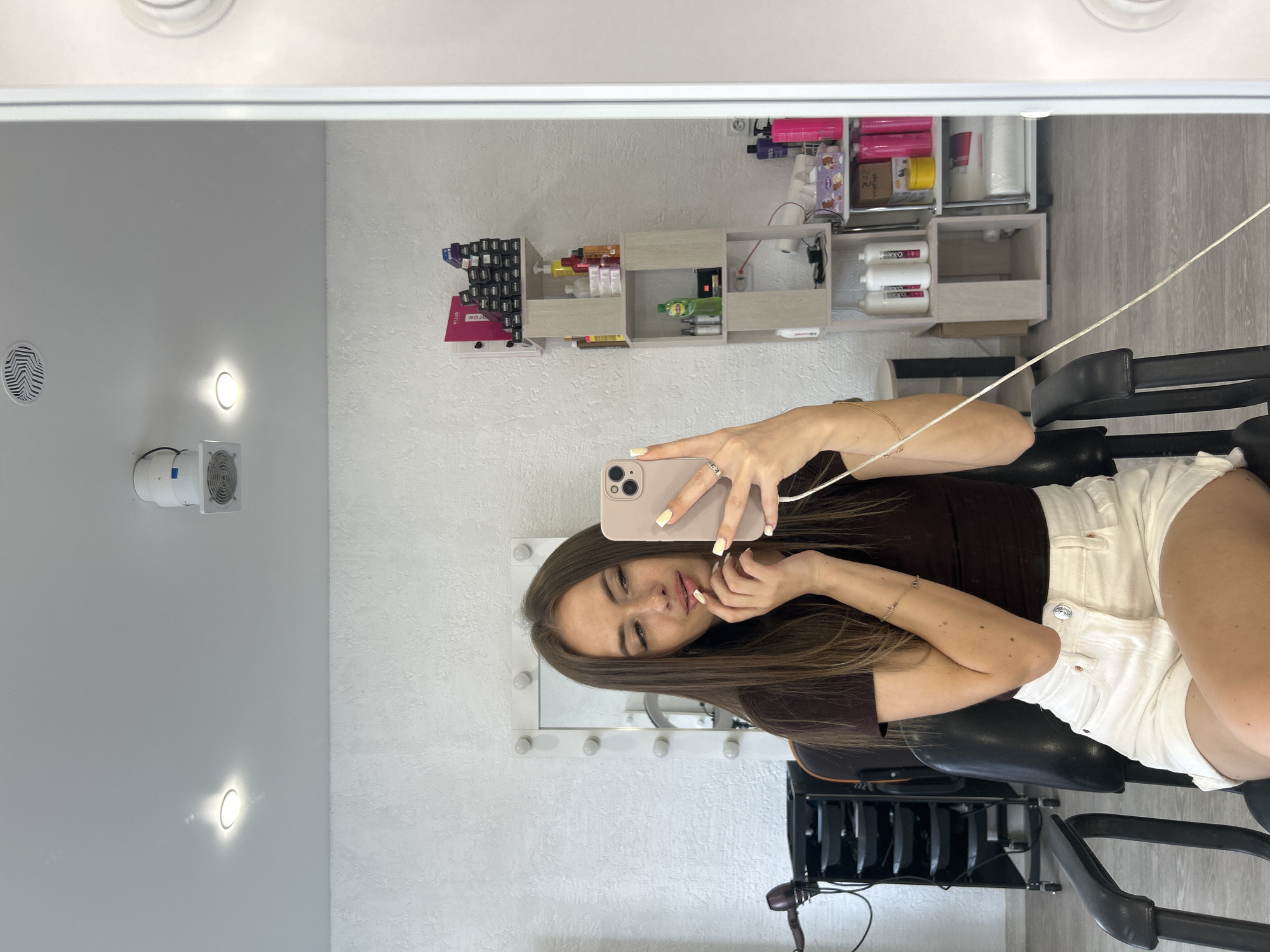 A young woman with long brown hair and a brown t-shirt and white shorts is sitting in a black salon chair and taking a selfie in a mirror. She is holding a pink smartphone in her right hand and her left hand is touching her face. Behind her, on the wall, is a large mirror with lights around its frame. To the left of the mirror is a black trolley with shelves, a hair dryer, and other salon equipment. To the right of the mirror, shelves are filled with various bottles and containers of hair products. The ceiling has a recessed light and a white cylindrical fan. The floor is gray.