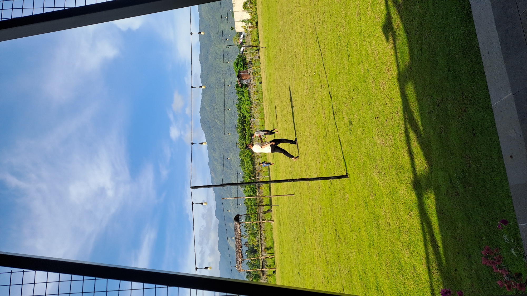 A wide shot captures an outdoor venue in Banda Aceh, Indonesia, with a vast, manicured green lawn under a bright blue sky dotted with wispy clouds. In the mid-ground, three individuals are scattered across the lawn. One person, wearing a white t-shirt and black pants, walks towards the right of the frame. Another person, further back and to the right, is also in motion, while a third, smaller figure is visible to the left of the walking person. Overhead, a string of bare light bulbs hangs horizontally across the scene, suspended by thin wires and supported by vertical poles. In the background, a line of lush green trees runs along the base of a range of rolling, verdant mountains that dominate the horizon. A simple thatched-roof structure with wooden supports is visible on the left side of the field. The foreground features a shadowed area cast by an unseen structure, with a hint of purple flowers on the lower left. The overall impression is one of a serene, open recreational space.