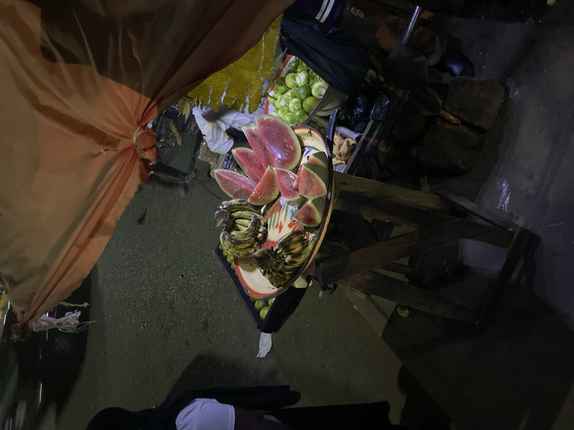This dimly lit, overhead view captures a vibrant fruit stall at an outdoor market, likely in Bukuru, Nigeria, at night. The central focus is a display of fresh produce on a wooden market stand. Slices of bright red watermelon, some still with rind attached, are neatly arranged on a colorful, patterned platter, reflecting the artificial light that illuminates the stall. Below the watermelon, several bunches of ripe bananas, showing both green and yellow patches, are visible. To the left and behind the main fruit display, a bin holds a large quantity of small, round green fruits, possibly limes or another type of citrus.
The stall itself is constructed with wooden planks, and an orange-brown fabric canopy or tarp is tied back, partially visible in the upper left, providing shelter. The ground beneath and around the stall appears dark and somewhat damp, possibly asphalt or packed earth. In the deeper background, obscured by shadows, other market elements or debris can be vaguely discerned. A person’s lower body, clad in white and black clothing, is partially visible in the bottom right corner of the frame, indicating human presence. The overall ambiance is that of a bustling night market, where the produce is strategically lit to attract customers. No text is visible in the image.