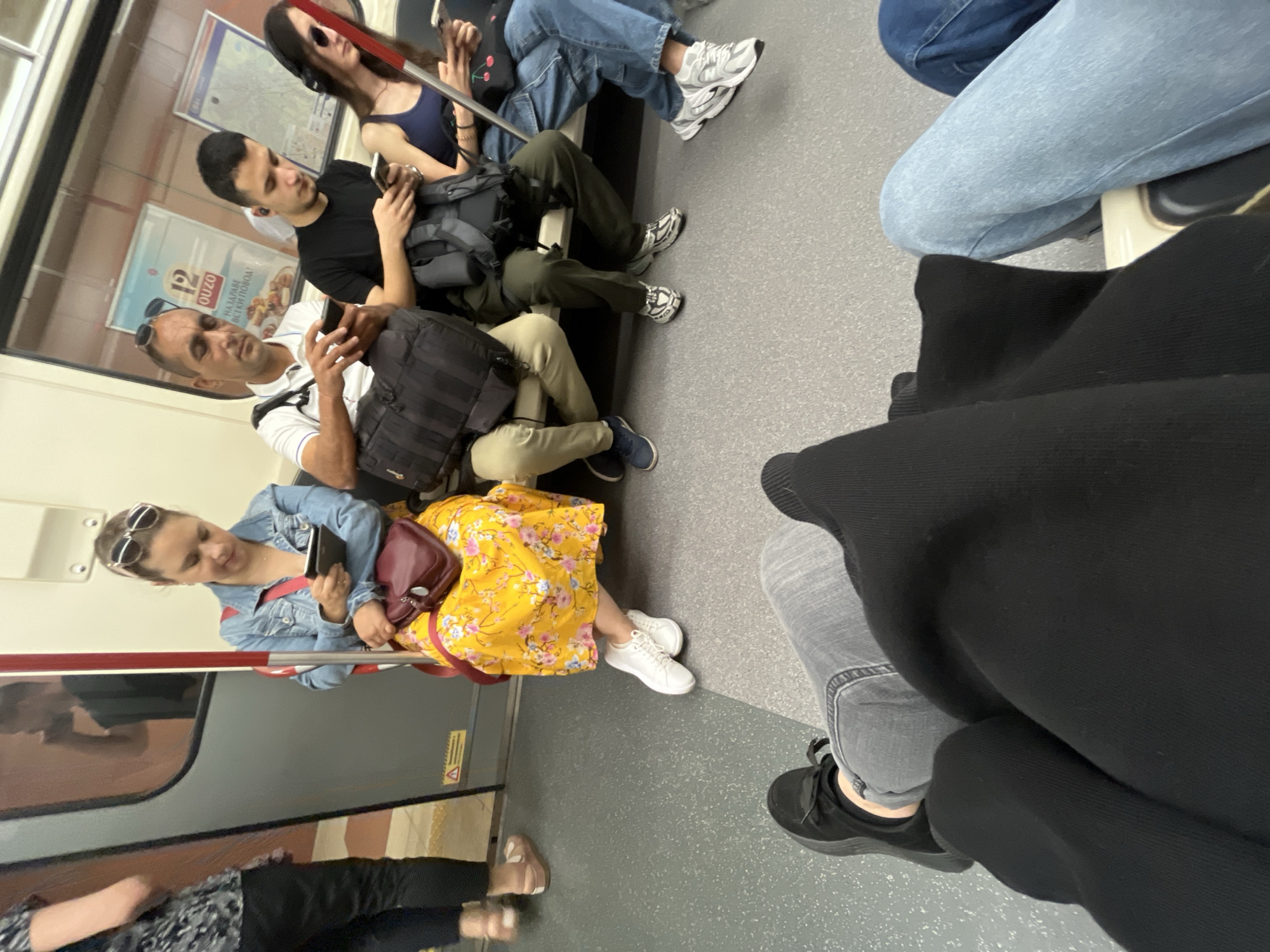 The image depicts the interior of a Sofia Metro train car. Several passengers are seated on a bench. In the foreground, the legs of two individuals are visible, one wearing black sneakers and grey jeans, the other in a black garment. To the left, a person in a black and white patterned top and black pants stands. A woman with sunglasses on her head, wearing a denim jacket and a bright yellow floral dress, is seated and looking at her phone. Next to her, a man in a white polo shirt is also using his phone. To his right, a young man with headphones is looking at his phone. Further to the right, a woman in a dark blue tank top and denim jeans is also using her phone. The environment includes the train's interior features, such as windows with advertisements, a door, and the grey floor.
