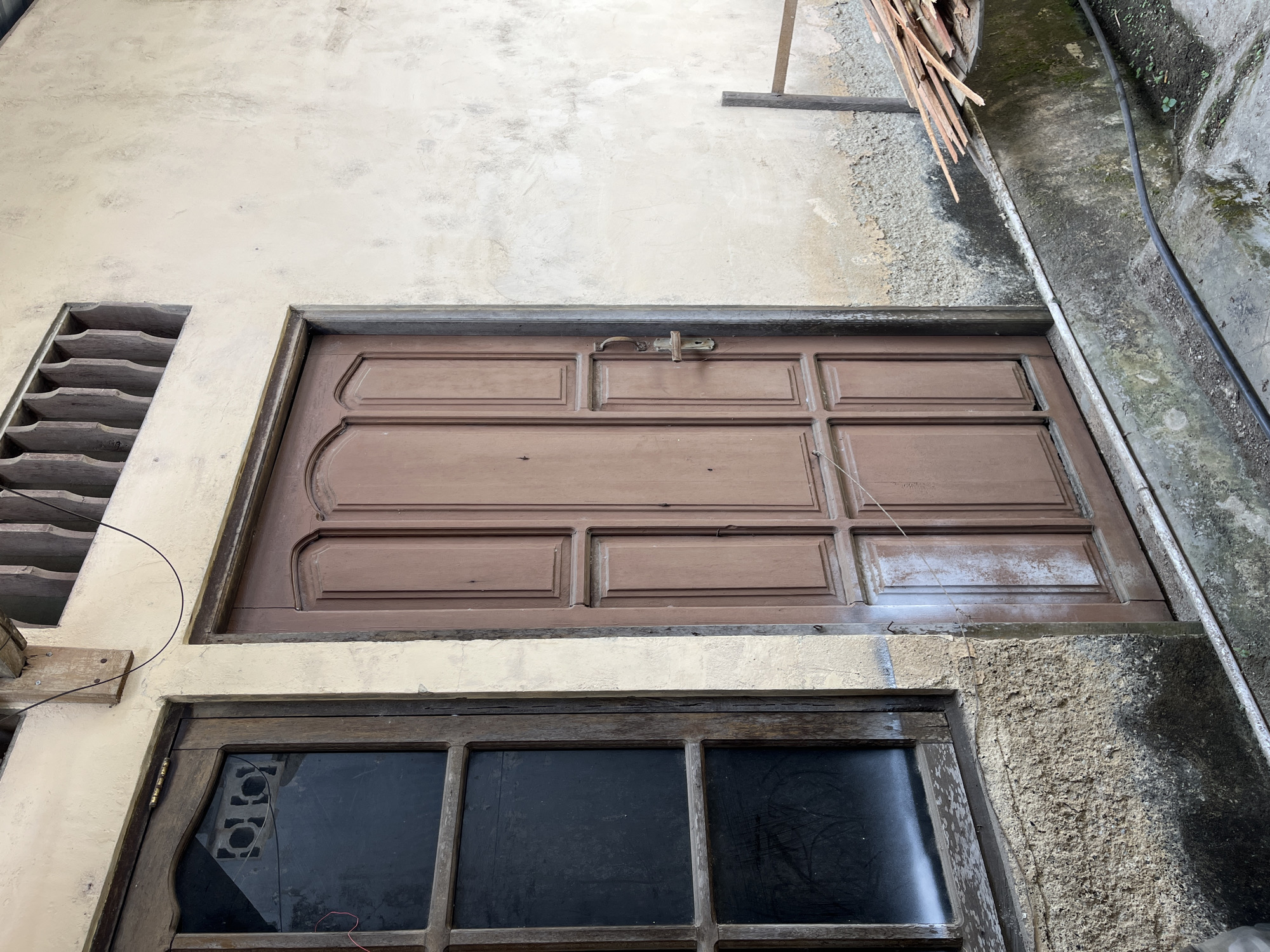 The image shows the exterior of a building in Sejahtera, Indonesia, featuring a wooden door and a window. The door is brown and has a traditional design with multiple panels. A metal door handle is visible on the right side of the door. To the left of the door is a wooden-framed window with dark glass panes. The wall of the building is textured and appears to be made of concrete or stucco, with some signs of wear and discoloration. Above the door and window, a wooden decorative element with vertical slats is present. In the lower right portion of the image, a pile of wood planks is stacked on a platform, and a black cable runs along the ground. The overall scene suggests a rustic or older building in a residential or commercial area.