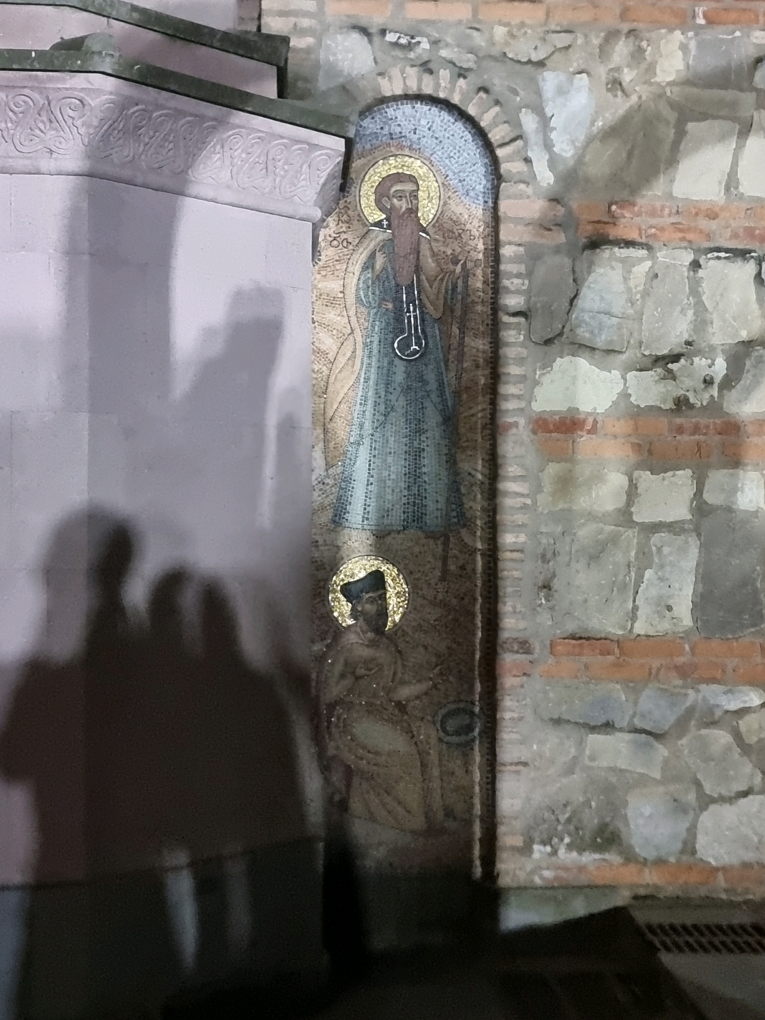 An outdoor, eye-level, medium shot captures a mosaic niche embedded in a rough stone and brick wall. The niche contains two religious figures. The upper figure, larger in scale, depicts a bearded man in a blue robe and a golden halo, holding a staff with a looped top. The lower figure, smaller, shows a man in a brown robe and a dark hat, also with a golden halo, holding a circular object. The wall surrounding the niche is constructed of irregular grey stones interspersed with reddish-brown bricks. To the left of the niche, a large, light-colored, fluted pillar is visible, casting dark shadows of three people onto its surface. The lighting is dim, suggesting nighttime or an indoor setting with limited illumination. A dark, indistinct object, possibly a grate or drain, is visible in the lower right corner.