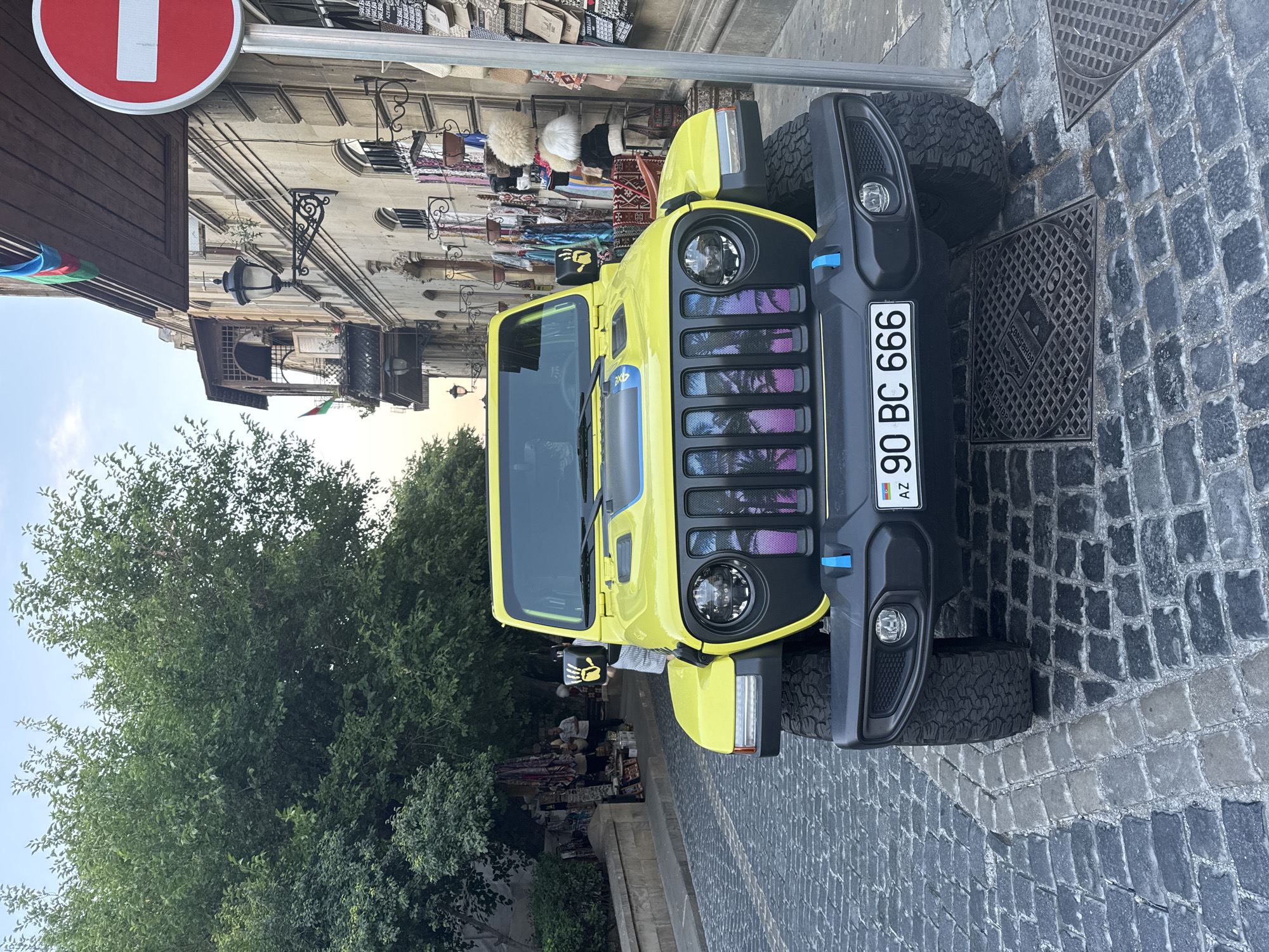 A bright yellow Jeep Wrangler is parked on a cobblestone street in Sabail Raion, Azerbaijan. The Jeep has a custom grill with a tropical palm tree design that is illuminated with purple and pink lights. The license plate reads "90 BC 666 AZ". In the background, there is a building with balconies and shops selling various items, including carpets and hats. A red and white "no entry" sign is visible on the right side of the image. Trees are on the left side of the image. The sky is overcast.