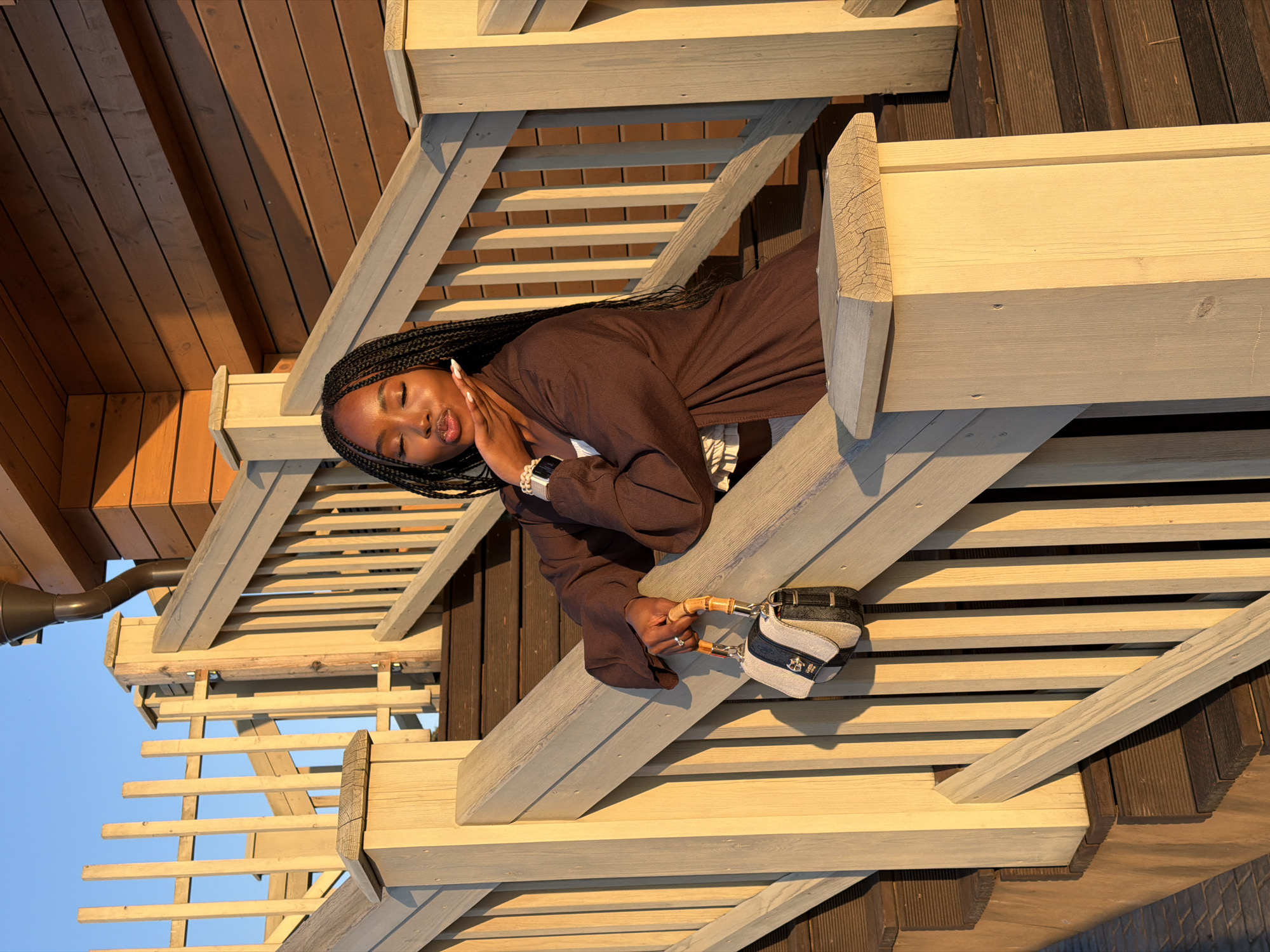 A young Black woman with braided hair is posing on wooden stairs during sunset. She is wearing a brown open-front jacket over a white top and is holding a small handbag with a bamboo handle. The stairs are made of light-colored wood and are part of a building with wooden siding. The sun is setting, casting long shadows and creating a warm, golden light. The location is the city of Kazan, Russia.
