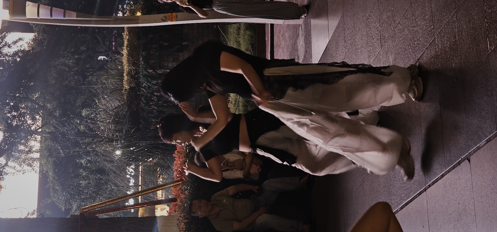 Two women are interacting, possibly dancing or adjusting each other's clothing, in what appears to be an outdoor urban environment. They are dressed in clothing that includes dark tops and light-colored, flowing bottoms. The location features trees, lighting fixtures, and paved surfaces, which suggest a park or public space. Other people are nearby, including one man reclining and another standing. The general location, based on the prompt, is Liangjiang, China.
