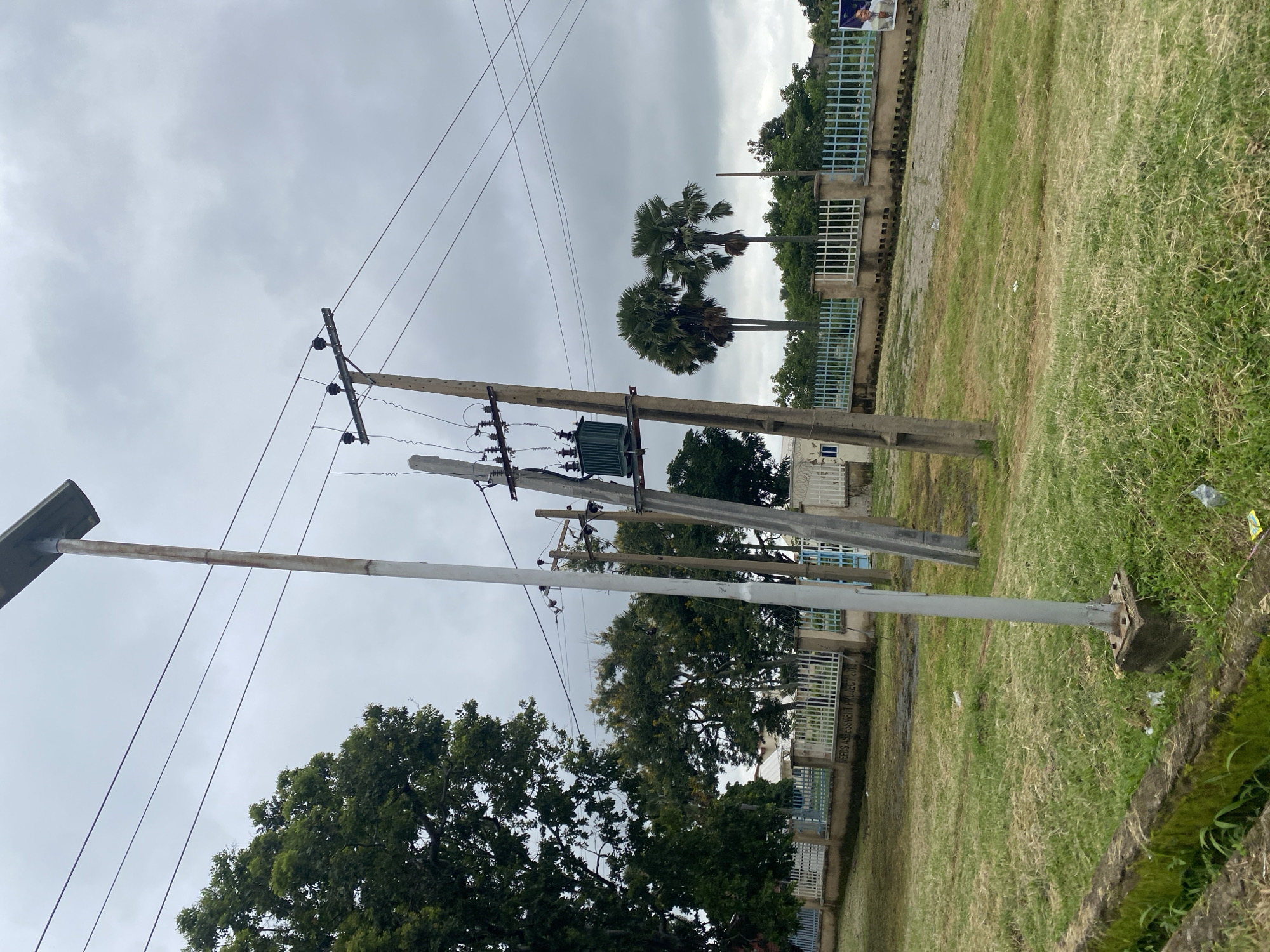 The image depicts an outdoor scene with power lines and utility poles in Yelwa, Nigeria. In the foreground, there is a grass-covered area with a concrete border. A tall, slender metal pole with a solar panel at the top stands to the left of the frame. To its right, a concrete utility pole supports electrical wires and a green transformer. Behind these structures, a light blue metal fence runs horizontally across the midground, separating the grassy area from a paved path and a building complex. Several trees, including palm trees, are visible in the background, under a cloudy sky. A poster featuring a person is affixed to a wall on the far right side of the image. The overall environment appears to be a somewhat overgrown, semi-urban area.