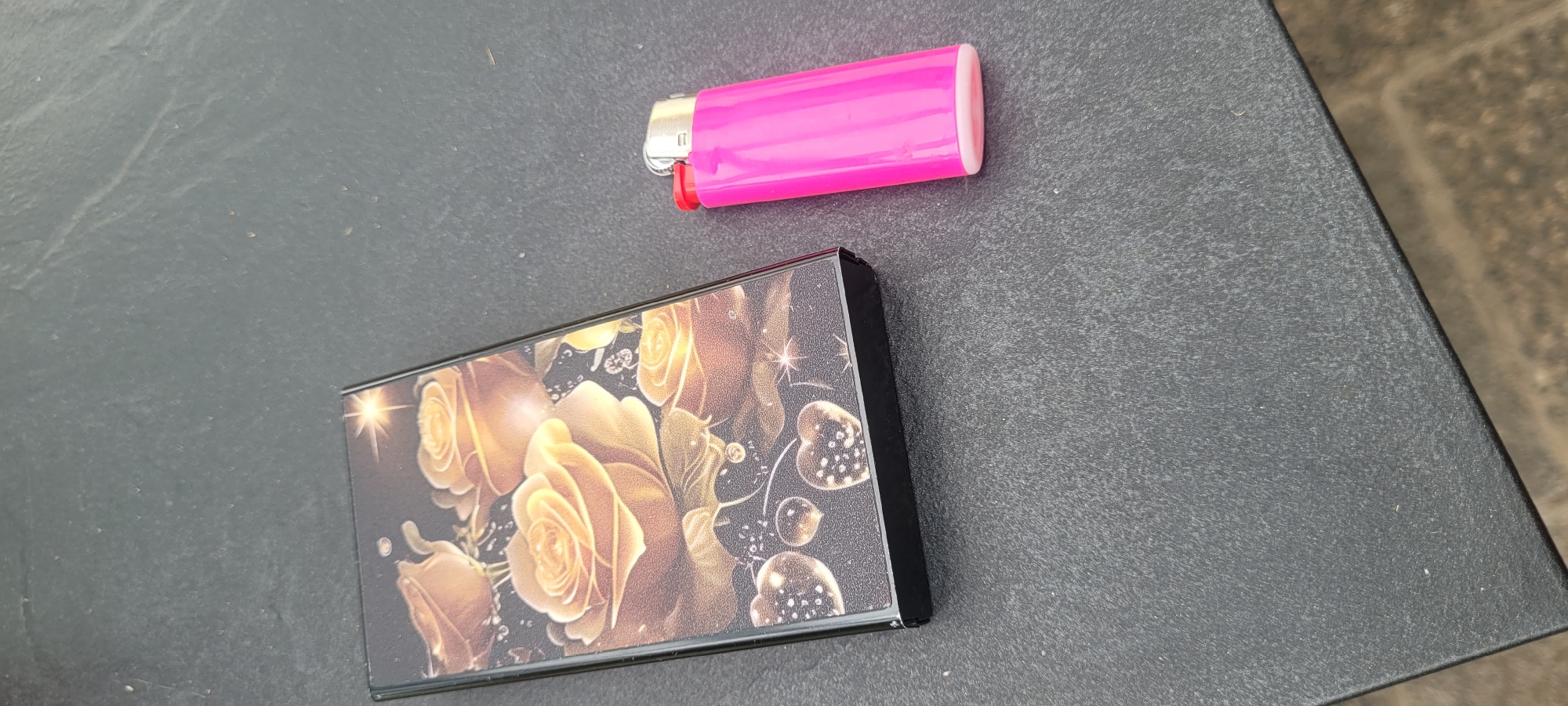 On a dark, textured outdoor surface, likely a table, two objects are visible: a cigarette case and a pink lighter. The cigarette case is rectangular and decorated with an intricate pattern of golden roses and glittering hearts. To its right, a bright pink lighter with a silver and red top is placed. The background is out of focus, suggesting an outdoor setting.