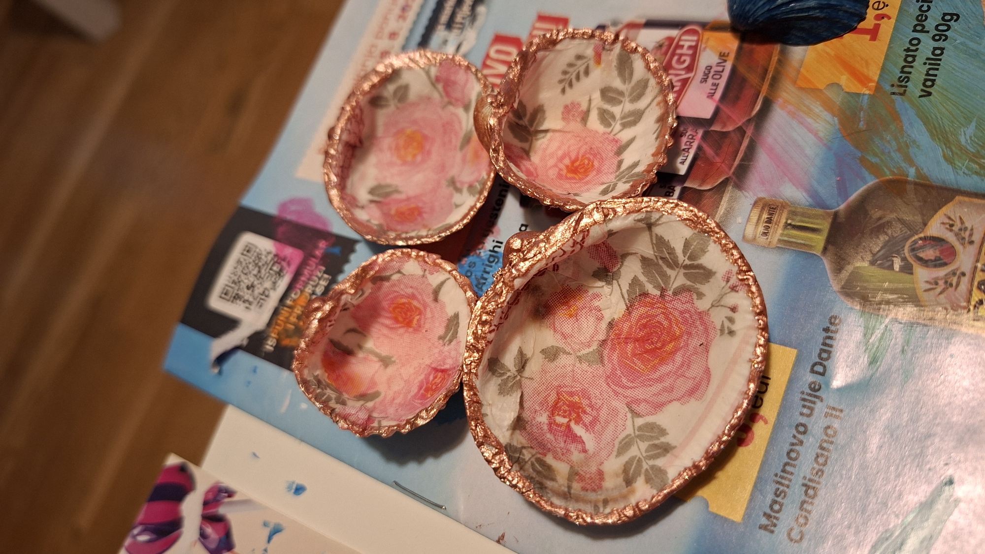 The image shows four decorative seashells arranged on a colorful, glossy surface, likely a magazine or brochure. Each seashell is lined with a floral patterned paper, featuring pink and red roses with green leaves. The edges of the seashells are trimmed with a metallic copper-colored material. In the background, various text and images are visible, including what appears to be a QR code, product labels with text such as "Maslinovo ulje Dante Condisano II", "OLIO DANTE", "SUGO ALLE OLIVE", and "Lisnato pecivo vanila 90g". The overall setting appears to be an indoor environment, with a wooden surface visible in the upper portion of the image. There is no indication of Podgorica, Montenegro within the image.