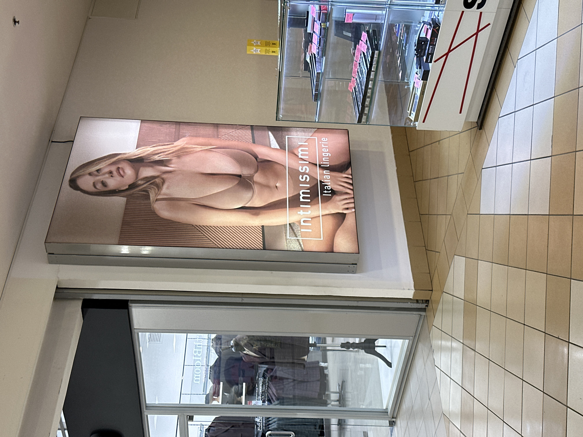 The image shows a store in an unidentified shopping mall. The store appears to sell clothing, as evidenced by the clothing racks visible through the glass storefront. Above the storefront is a large advertisement featuring a lingerie model and the brand name "Intimissimi". To the right of the advertisement is a glass display case containing various items, possibly cosmetics or accessories. The floor is tiled with beige and white tiles, and the lighting appears to be bright and artificial. The precise location cannot be determined from the image alone.
