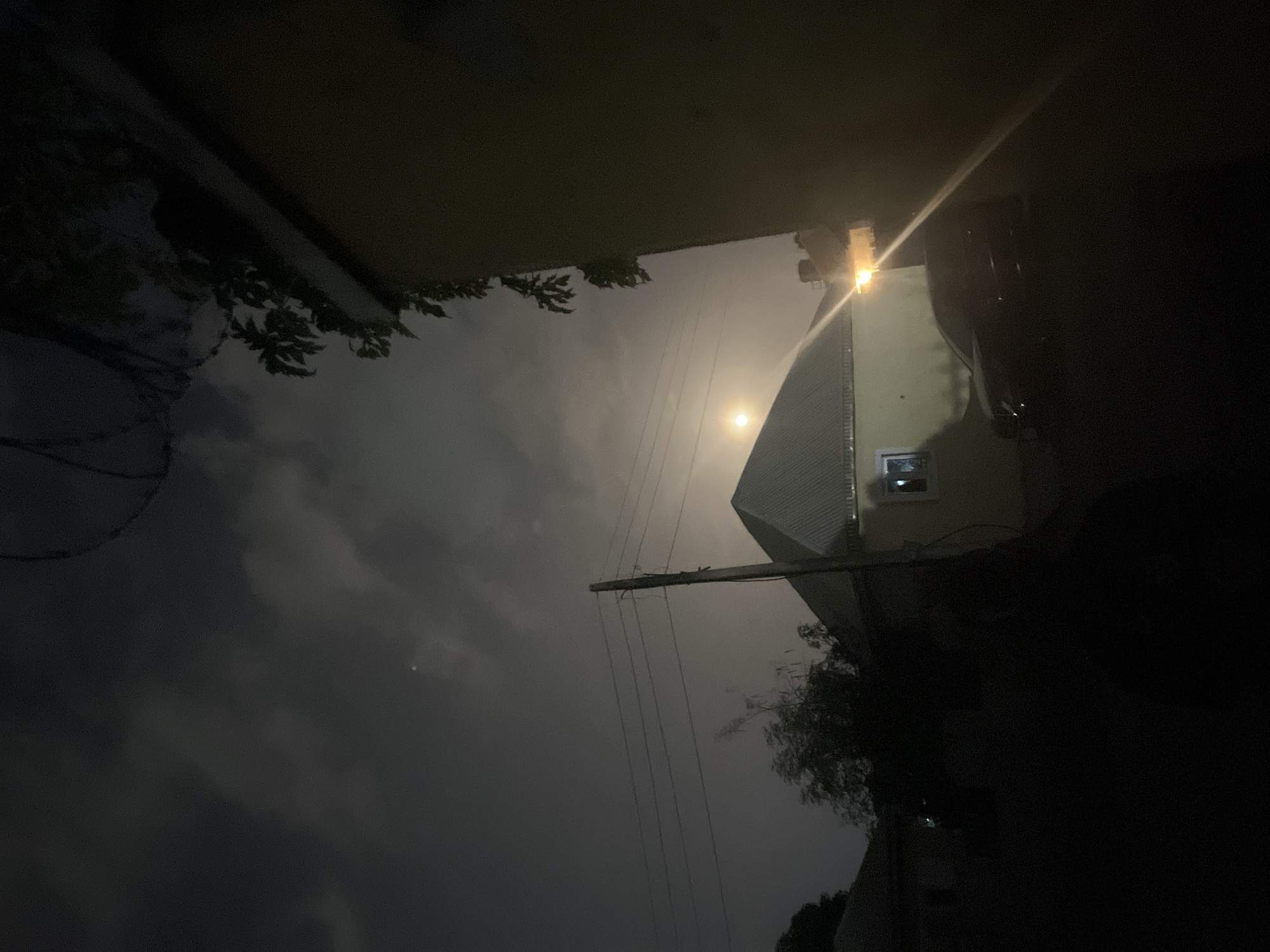 A low-angle nighttime view, tilted slightly, of a building with a pitched roof, illuminated by a streetlight. A moon is visible in the dark, cloudy sky. Power lines run diagonally across the frame. Overhanging foliage is present in the upper and lower left corners. Barbed wire is visible in the left midground. The location appears to be an urban or suburban area, potentially Abuja, Nigeria.
