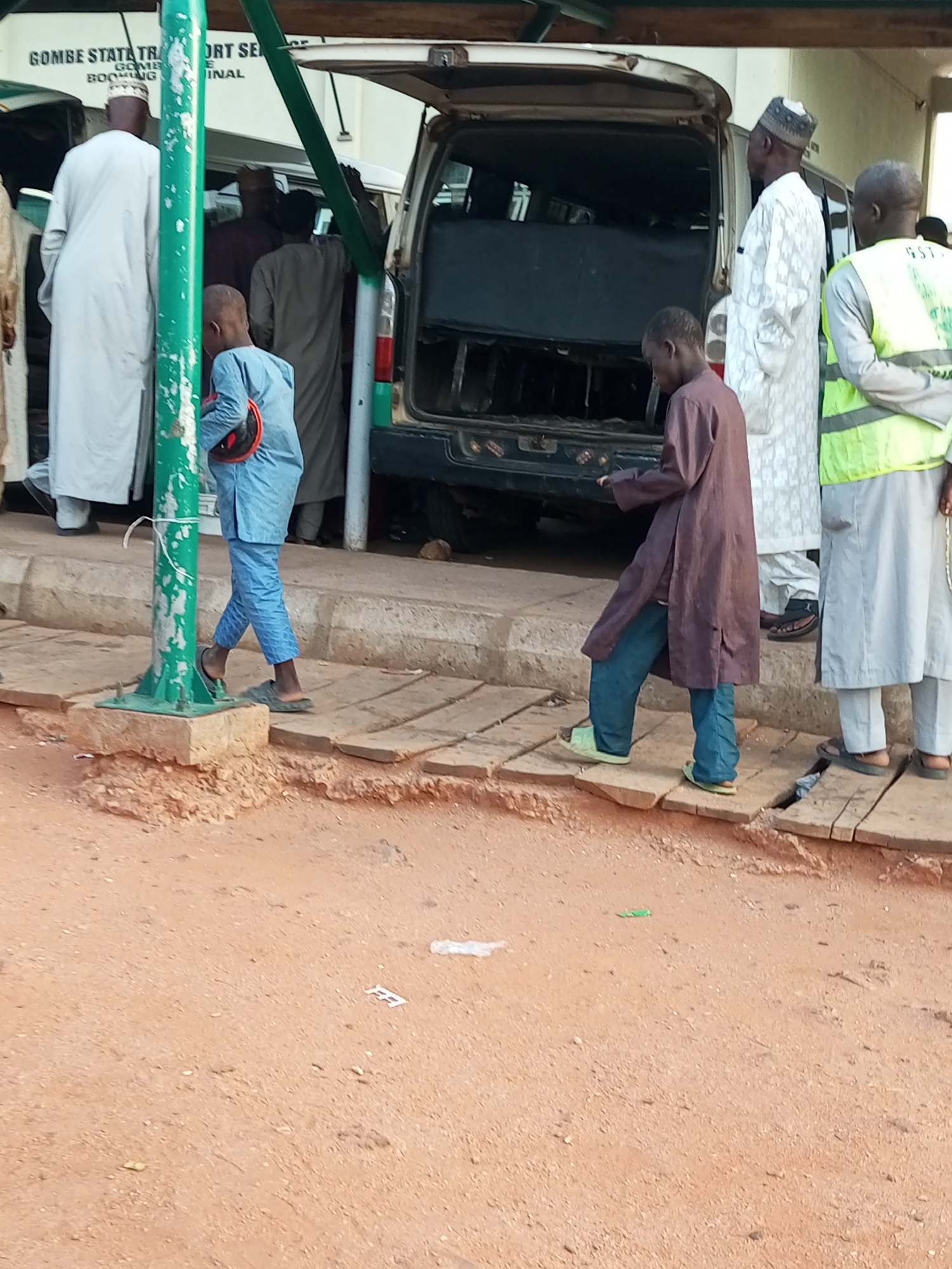The image depicts a scene at the Gombe State Transport Service booking office in Gombe, Nigeria. Several individuals are present, some appearing to be passengers and others potentially staff. In the foreground, a young boy in a light blue tunic and trousers, holding a red and black object, walks on a wooden walkway next to a green metal pole. To his right, a man in a dark brown robe and blue jeans walks away from the camera, also on the wooden walkway. Behind them, several other people are visible near a van with its rear doors open. One man in a white patterned robe and cap stands by the van, while another man in a bright yellow vest stands to the right. The environment appears to be an outdoor public transportation hub, with unpaved ground and a concrete curb visible.