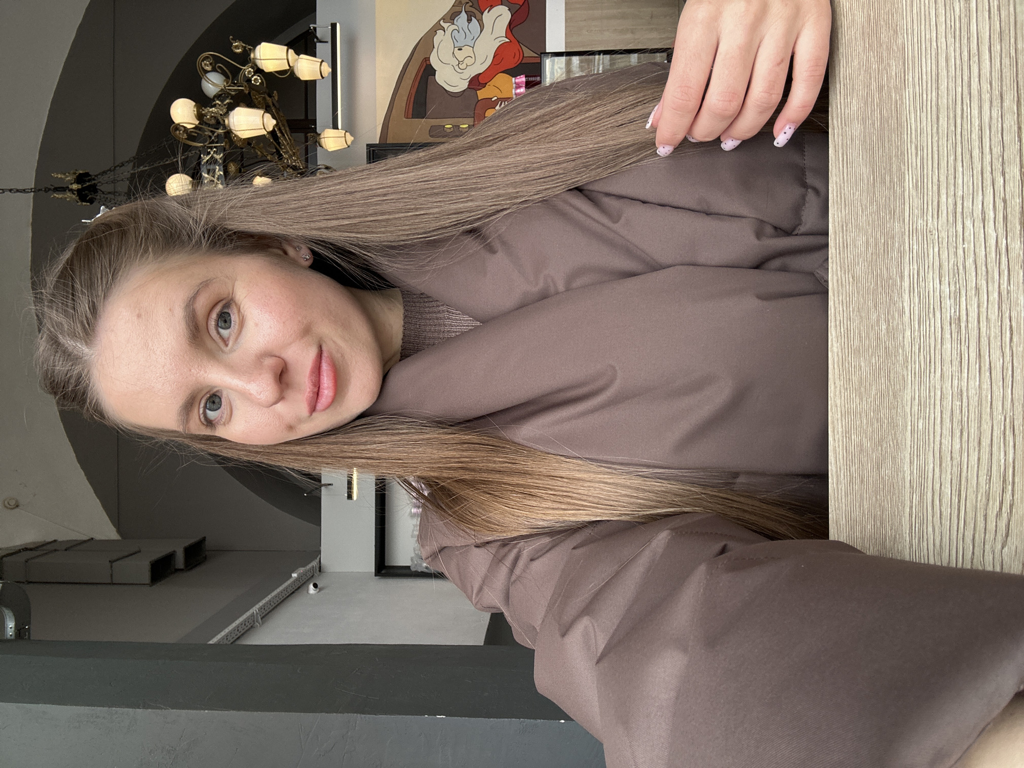 A young woman is captured in a selfie-style portrait, looking directly at the camera with a calm and subtly smiling expression. Her head is tilted slightly, and her long, straight, light brown hair cascades over her shoulder and chest. She is wearing a warm, brown puffer jacket over a lighter brown knitted top. Her right hand, featuring manicured nails with a light pink polish and small white polka dots, is visible near her hair.
The background suggests an indoor setting, likely a cafe or restaurant in Moscow, Russia. To the left and behind her, a large, ornate golden chandelier with multiple illuminated lamps hangs from the ceiling. Further back and to the right, a colorful wall mural is visible, depicting a cartoon-style character with a white beard and a red hat, reminiscent of Snow White's Dopey. A light-colored wooden table or counter occupies the right side of the frame, running vertically. The overall lighting is soft and ambient. There is no visible text in the image.