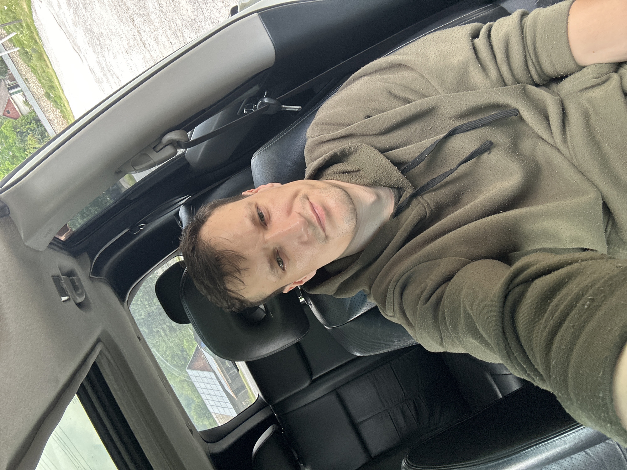 A Caucasian male is seen from the chest up, seated in the driver's seat of a vehicle. He is wearing a hooded sweatshirt. The vehicle's interior is visible, including black leather seats, a sunroof, and a seatbelt. Outside the vehicle, a rural landscape is visible with buildings, trees, and a gravel road. The image appears to have been taken in Makiv, Ukraine.