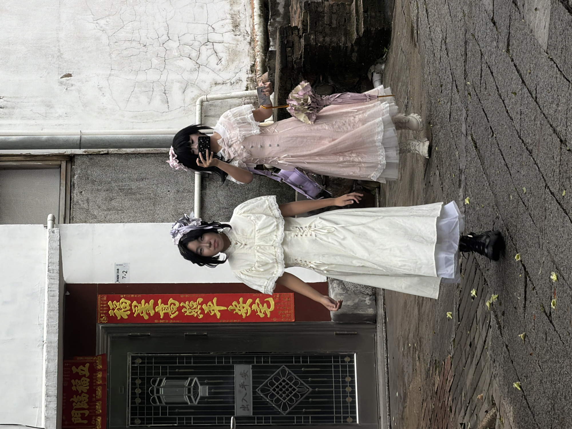 The image captures two young individuals walking on a cobbled street in front of a building with Chinese characters on a red banner and a dark-colored door with decorative metalwork. One individual, on the left, wears a white lolita-style dress with a lace collar and corset-like lacing at the waist, black boots, and a headpiece. This individual is looking forward. The other individual, on the right, wears a pink lolita-style dress with a white lace overlay, white boots, and a pink floral hair accessory. This person is holding a phone to their face, appearing to take a photo, and carries a light purple bag and a pink umbrella. The building appears to be in a somewhat aged urban environment, suggested by the cracked white walls and weathered stairs in the background. The presence of Chinese characters on the banner and the building's architectural style indicate an East Asian location, potentially in the city of Longgang District, China, given the provided information.