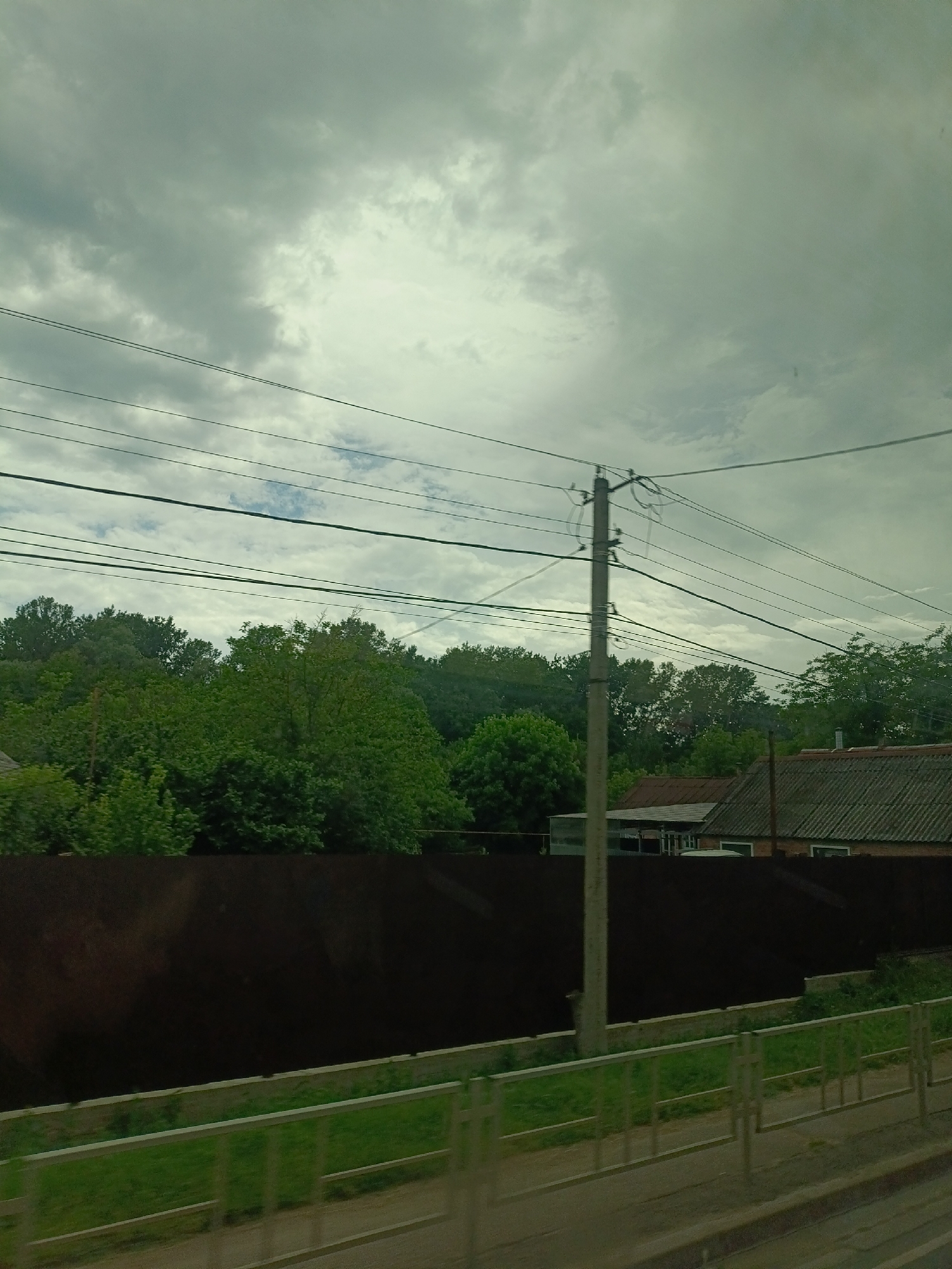 The image captures a view of a somewhat overcast sky with scattered clouds, over a residential area that appears to be in Saratovskaya, Russia. In the foreground, there's a metal fence with green grass behind it, and a dark, solid fence extending across the middle ground. Beyond the dark fence, a cluster of lush green trees forms a dense backdrop. To the right of the trees, the roofs of several houses are visible, characterized by their tiled or corrugated surfaces and chimneys. A concrete utility pole stands prominently in the mid-ground, supporting multiple overhead electrical wires that stretch across the frame. The overall impression is one of a suburban or semi-rural setting under a typical day's weather conditions.
