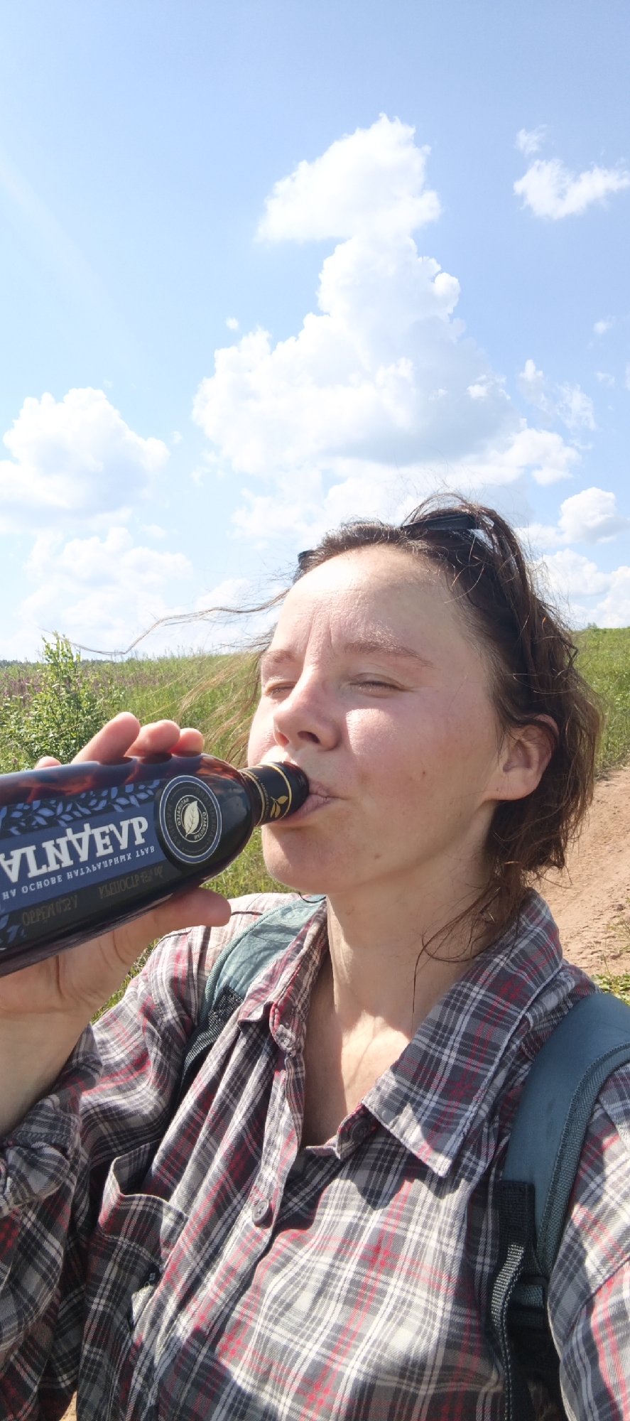 An outdoor, medium shot captures a woman drinking from a dark bottle. The woman is seen from the chest up, with her eyes closed and her head tilted back slightly. She is wearing a plaid shirt and a blue backpack. The bottle she is drinking from has a label with text that is partially visible. The background is a grassy field under a bright blue sky with white clouds. The lighting suggests it is daytime. The image was taken in the vicinity of Bol'shoi Bukor, Russia.