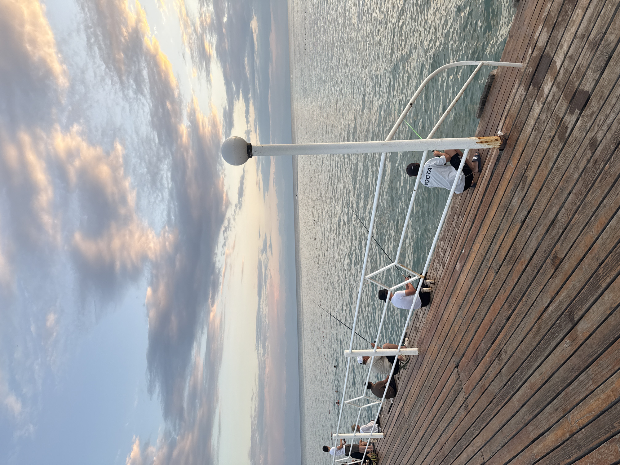 The image shows a wooden pier on a body of water, likely Issyk-Kul Lake in Sary-Oy, Kyrgyzstan, during what appears to be late afternoon or early evening given the soft lighting and clouds. Several people are present on the pier, engaged in fishing. In the foreground, the weathered wooden planks of the pier stretch out, with a white railing running along the edge. A white lamp post with a spherical top stands on the right side of the pier.
From left to right, there are at least six individuals visible. The person furthest left is standing and appears to be holding a fishing rod. Next, there are two people seated, one in a white t-shirt and hat, and another in a brown shirt. Further along, a man in a beige t-shirt and white hat is seated on a small stool, also fishing. To his right, another person in a white t-shirt is seated, holding a fishing rod. The person furthest to the right is wearing a grey t-shirt with "NOCTA" printed on the back and black shorts, also fishing.
In the water behind the pier, several buoys are visible, and a few people can be seen swimming or wading. The water is calm with gentle ripples, reflecting the pastel colors of the sky, which is filled with soft, wispy clouds tinged with pink and orange. The horizon shows a faint, hazy outline of distant landmasses or mountains. The overall atmosphere is serene and recreational.