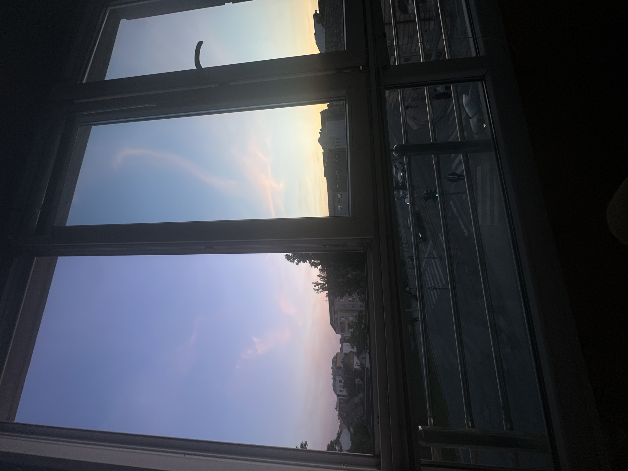 The image shows a view from a window in Novi Sad, Serbia, overlooking a cityscape at dusk. Three window panes offer different perspectives: the top pane reveals a sky with yellow hues, suggesting either sunrise or sunset; the middle pane shows a slightly lower angle of the same sky, capturing buildings in silhouette; the bottom pane presents a view towards residential buildings under a darkening sky with pink tones. To the right of the window, visible through a barred section, is a street with cars and pedestrian crossings. The room is dimly lit, creating a contrast with the outside light.
