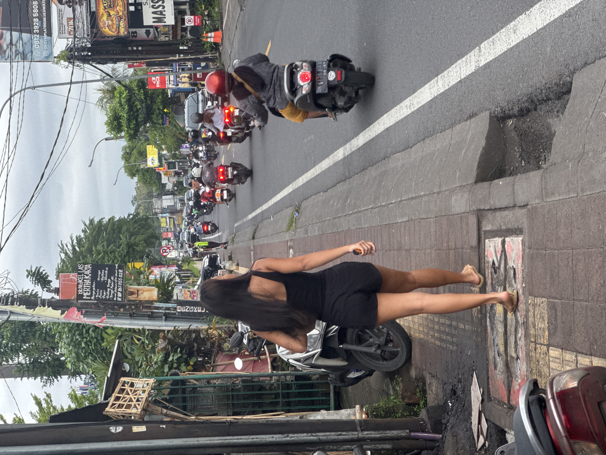 This image captures a bustling street scene in Kuta Selatan, Indonesia, during what appears to be an overcast day. In the foreground, a woman with long dark hair, wearing a black strapless romper or dress and sandals, stands on a tiled sidewalk, observed from behind. She is positioned beside a white and black scooter parked on the sidewalk. Her left hand holds a small, light-colored object.
The street, adjacent to the sidewalk, is characterized by heavy traffic, predominantly composed of motorcycles, some of which appear to be stopped. One rider in the mid-ground is identifiable by a red helmet and a dark jacket over yellow shorts, with their motorcycle displaying the license plate "DK 4136 PAO." A distinct white line marks the edge of the paved road.
The background features commercial buildings and lush green foliage, interspersed with numerous utility poles supporting a dense network of overhead wires. Various street signs and billboards line the thoroughfare. A broken concrete curb with damaged pavement is visible between the road and sidewalk. Further down the sidewalk, a woven wooden cage-like structure rests on a stand.
Several pieces of text are visible on the signs: "BENGKEL LAS MERTHA KARYA" (Welding Workshop Mertha Karya) with a list of services including "Tralis, Hanopi, Pintu, Balkon, Pagar, Stainless, Reiling Tangga, Tangga Putar.dll." (Trellis, Canopy, Doors, Balconies, Fences, Stainless, Stair Railings, Spiral Stairs, etc.) and a phone number "HP. 087 861 055 028." Another sign reads "DEGYMPLATINUM.ULUWATU" with the number "0812 3928 5808" and "70K." Portions of other signs, including "MASS," "S EAN S," "U," and "VP MOT," are also discernible. The scene presents a typical urban environment in Southeast Asia.