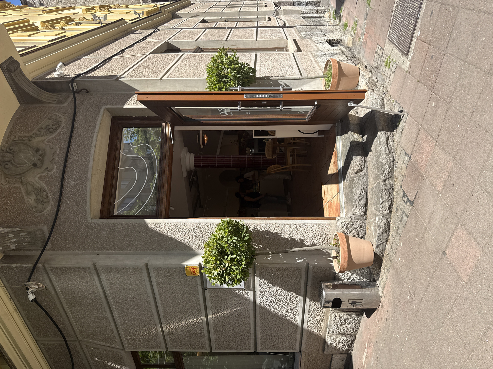 A street-level view showcases an open storefront in Belgrade, Serbia. The building's facade is constructed of light-colored stone, textured with horizontal and vertical lines creating recessed panels. A rectangular window is positioned to the left of the open entrance, reflecting exterior objects on its surface.
Two potted plants are stationed above the door, one on each side. Underneath the right plant sits a metal trash receptacle. The sidewalk adjacent to the building is paved with red bricks.
Inside the store, a glimpse of a tiled wall and some furniture can be seen. People can be seen inside.
The perspective is slightly tilted, giving a bottom-up view of the building. The architecture above the storefront is visible, featuring a complex roofline and decorative elements.