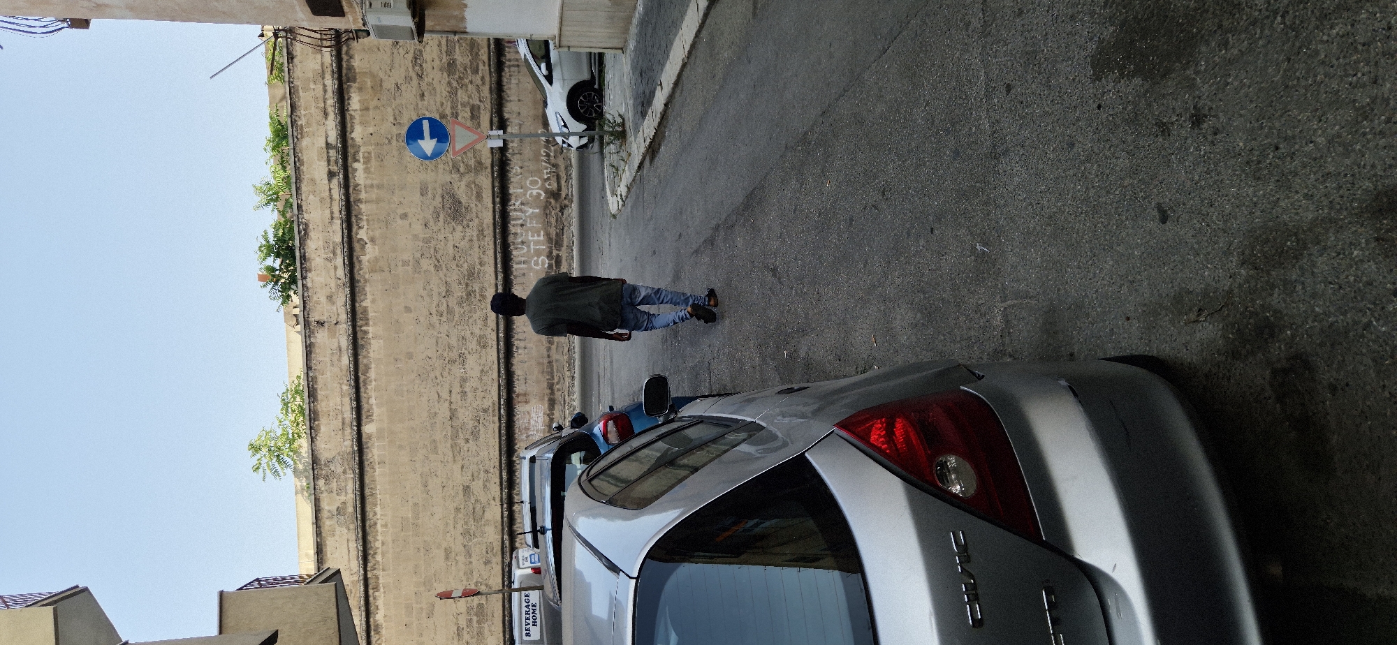 A lone individual, appearing to be a dark-skinned male wearing a dark green t-shirt and light blue jeans, walks away from the camera on a paved street. He is walking towards the right side of the frame. In the background, a large, weathered stone wall dominates the upper portion of the image. The wall has a rough texture and appears to be ancient. Above the wall, a clear blue sky is visible, with some greenery peeking over the top.
On the right side of the street, a white car is parked, and a white and blue traffic sign with a left-pointing arrow is mounted on a pole. Below the arrow sign is a red and white triangular sign. Graffiti is visible on the stone wall behind the signs and the white car, with the words "STEFY 30" clearly legible.
In the foreground, a silver car, identified by the "CIVIC" badge on its rear, is partially visible, taking up the left side of the frame. The car's rear left taillight is prominent. Several other cars are parked along the street on the left side of the frame, including a blue car. The street itself is asphalt, with some cracks and stains visible. The overall impression is an urban street scene in a historical area.
The scene is consistent with the architecture and street layout found in Taranto, Italy, particularly in areas with older fortifications and buildings. The stone wall is reminiscent of the historic city walls or ancient structures present in the region.