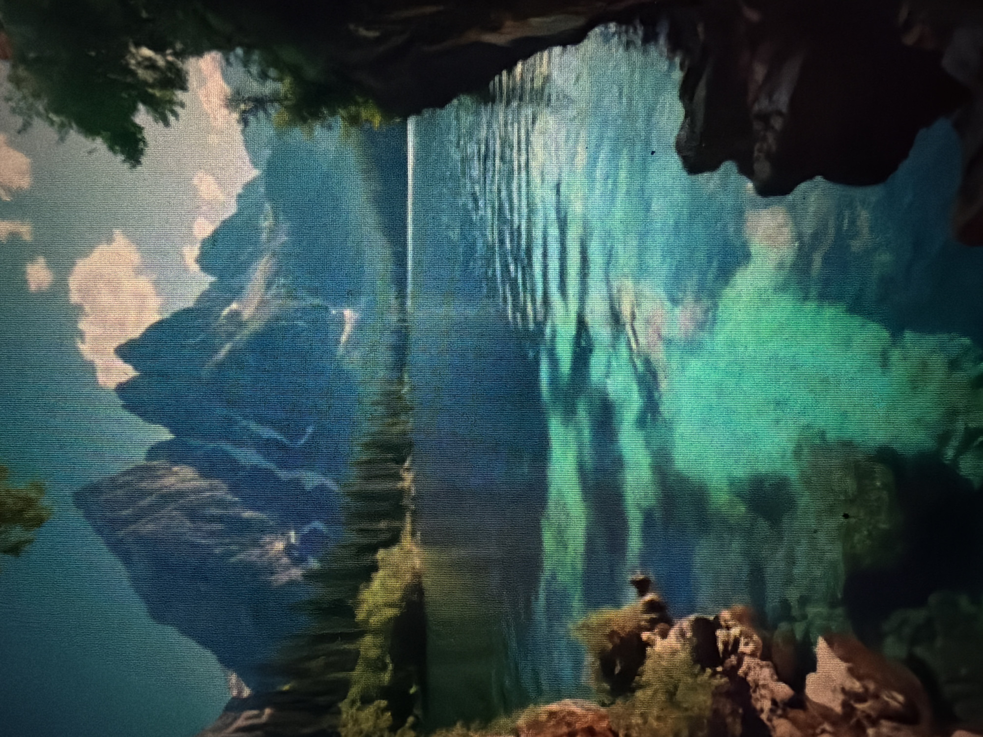 The image depicts a serene mountain lake scene. Jagged, majestic mountains rise in the background, their peaks partially obscured by soft clouds and bathed in a cool, blue light. A dense evergreen forest lines the shore, its dark green hues contrasting with the lighter green of the trees closer to the water. The lake itself is a striking turquoise, so clear that the sandy bottom and submerged rocks are visible beneath the rippling surface. The water is calm, with gentle waves reflecting the sky and surrounding landscape. On the right side of the frame, dark, rocky cliffs descend towards the water, framing the view. The overall atmosphere is one of tranquility and natural beauty. The image is not set in the city of Baan Mae Nam, Thailand, as it depicts a mountain and lake environment typical of alpine regions, not the tropical landscapes of Thailand.