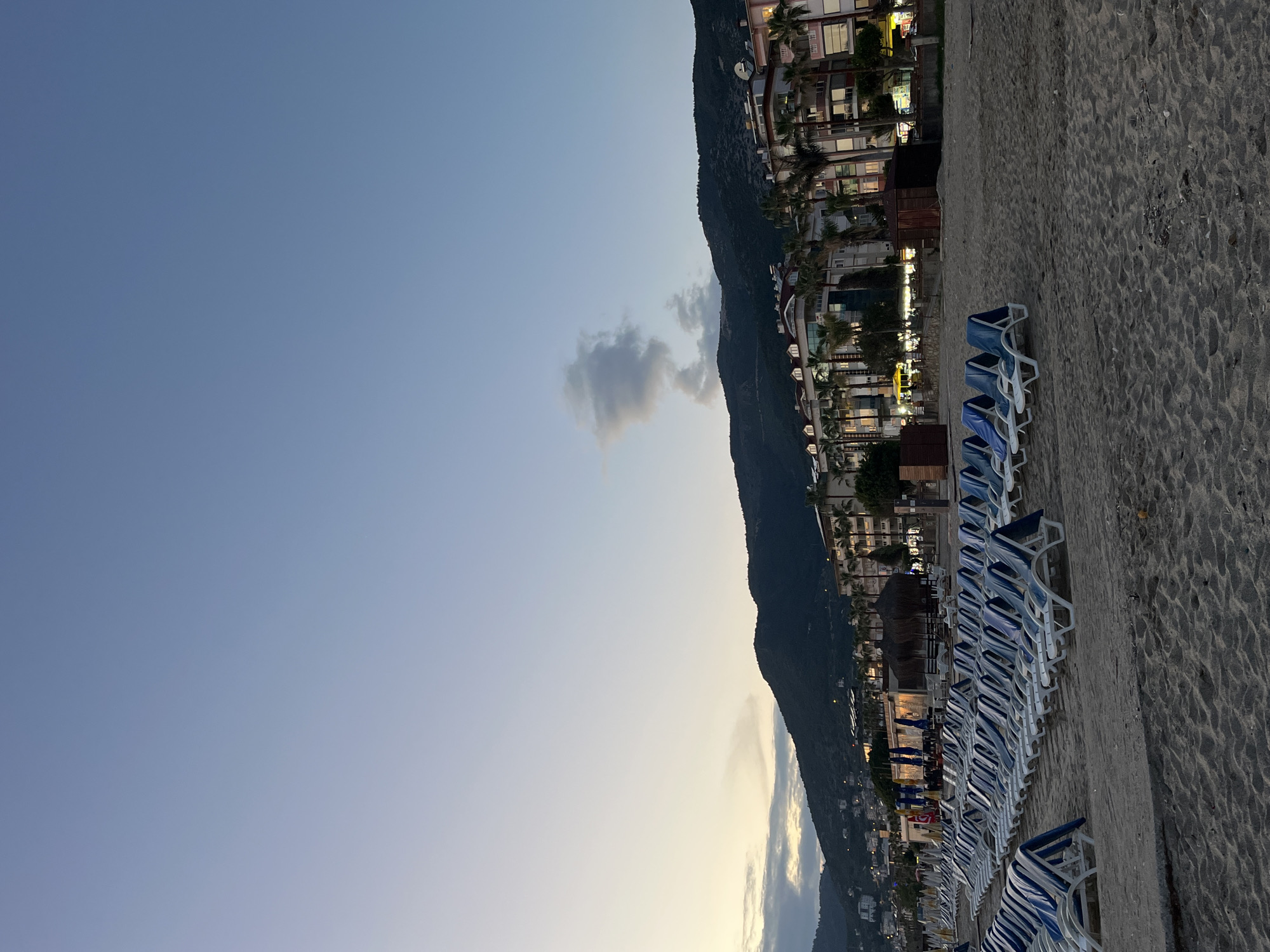 The image shows a beach scene in Alanya, Turkey at dusk. Rows of blue and white beach chairs line the sandy beach. A mountain rises sharply behind a series of buildings with lights on, suggesting they are hotels or residences. The sky is a gradient of blue to pale yellow near the horizon. There is a lone cloud in the sky.