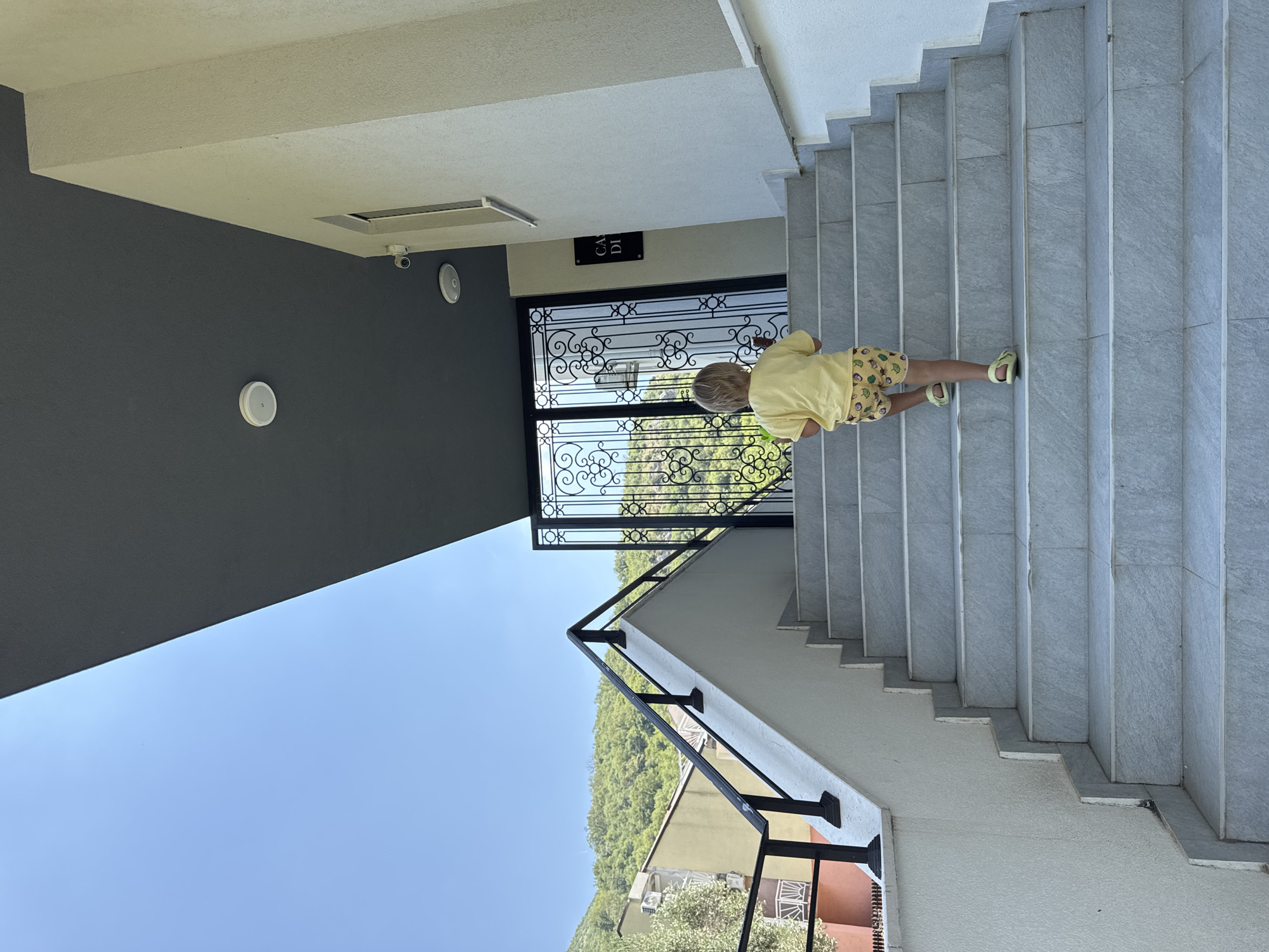 A young child, seen from behind, wearing a yellow t-shirt and patterned shorts, walks up a set of outdoor stairs. The stairs are made of light-colored stone with dark grey railings. Above the stairs, there is a dark grey wall with two white circular objects and a rectangular ventilation grate. Next to the ventilation grate, there is a black sign with white text. The scene is set in an outdoor environment with a clear blue sky and a background of green, tree-covered hills. The overall impression is of a sunny day in a residential area, possibly in the Mediterranean.