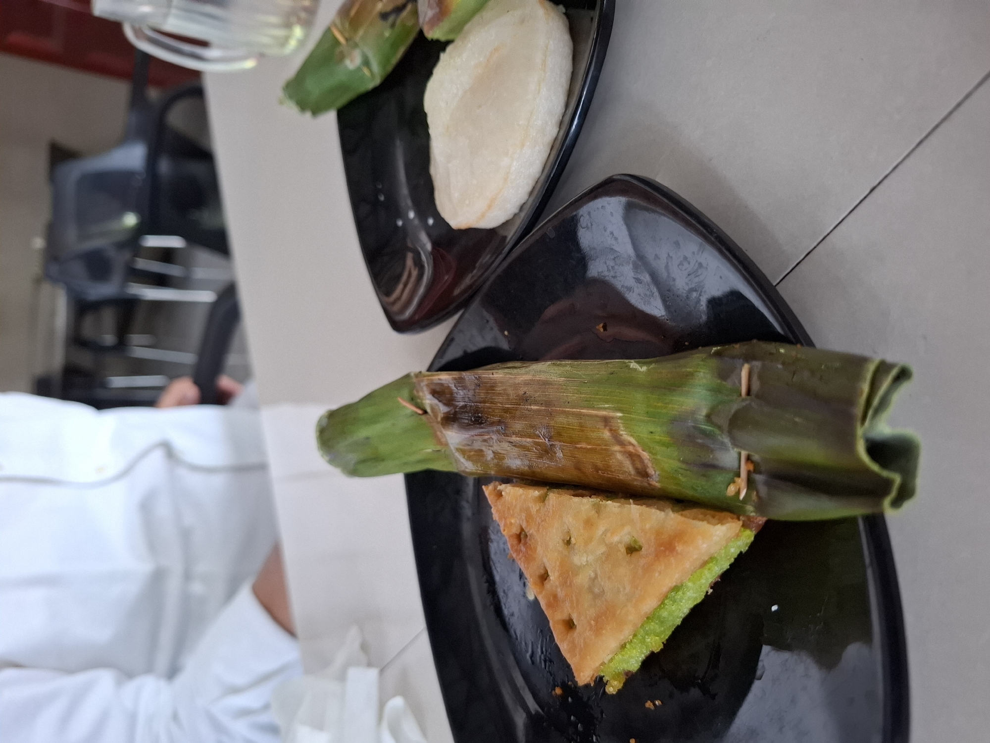 A high-angle shot depicts two black, triangular plates, each containing food. The plate closer to the camera holds a triangular piece of golden-brown food with a green layer at its base, partially covered by a green, cylindrical object wrapped in leaves and lightly scorched. The other plate holds a white, oval-shaped item, also partially covered by similar leaf-wrapped food. The scene suggests a meal, possibly consisting of traditional Indonesian snacks or desserts. The location is Lhokseumawe, Indonesia. A person wearing a white shirt is visible in the background. A black chair is also partially visible. The table is a light grey tile.